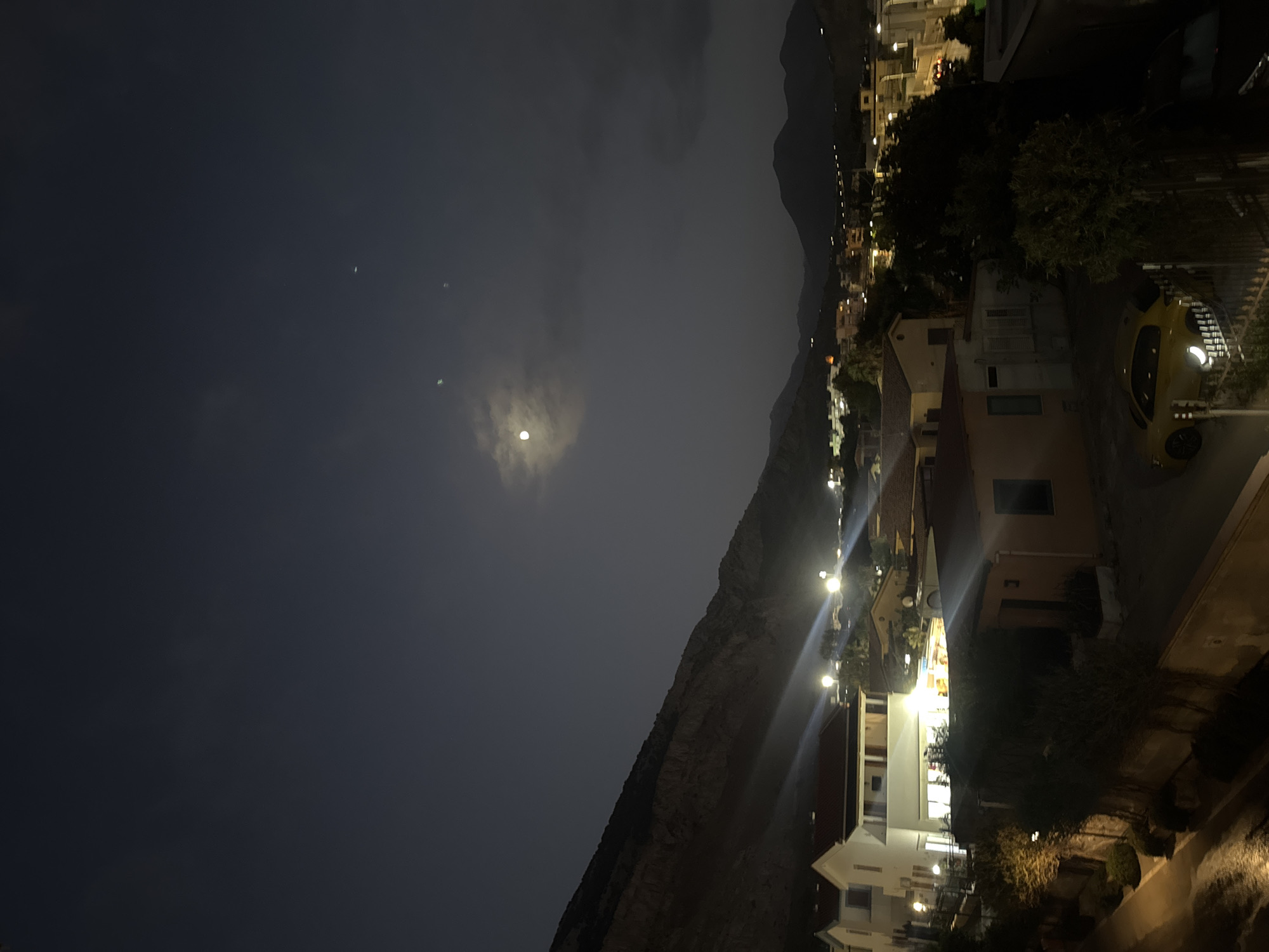 The image depicts a nighttime aerial view of Capaci, Italy, featuring a residential area nestled against a mountainous backdrop. The sky is dark with a visible moon and a few faint celestial bodies, suggesting it is likely evening or early morning. Streetlights and lights from buildings illuminate the scene, casting long beams across the landscape. In the foreground, several houses with tiled roofs are visible, some appearing modern with bright interior lighting. A yellow sports car is parked in front of one of the residences, its headlights on. Trees and vegetation are scattered throughout the scene, some of which are illuminated by the artificial lights. The overall impression is that of a quiet, moonlit night in a Mediterranean coastal town.