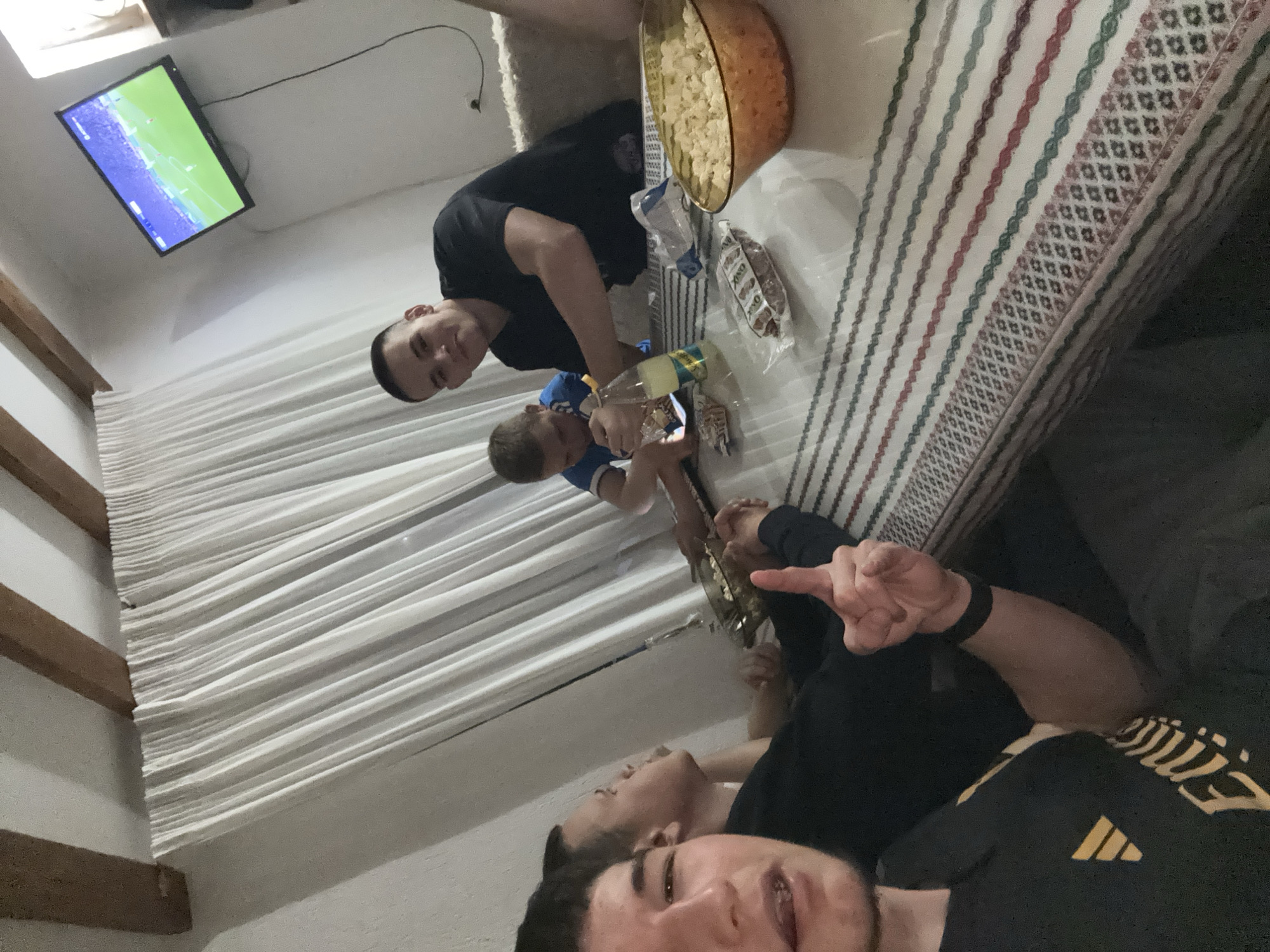 The image shows a group of four people in an indoor setting. One person, positioned at the bottom of the frame, is taking a selfie and has a black shirt with a yellow logo, a black wristband, and is pointing with his finger. To his left is another person in a black shirt who is lying down. Above these two, a person is standing and filling glasses with a liquid from a clear bottle. To the left of them is a boy in a blue shirt. Above them is another person standing and looking downward. A large bowl of popcorn and snack packages are present next to a striped couch or bed. In the background, a TV displays a soccer match. The room also features white curtain blinds. The location is unspecified but can be inferred that this is a residence in Zlatograd, Bulgaria.