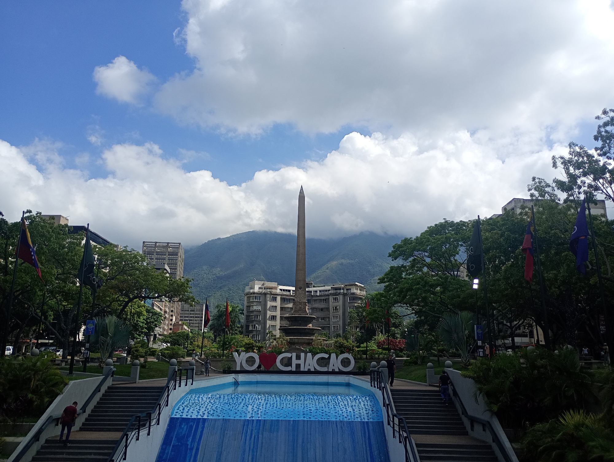 The image depicts a plaza in the Chacao Municipality of Caracas, Venezuela. A large obelisk monument stands centered in the background, surrounded by buildings and a backdrop of mountains partially obscured by clouds. In the foreground, a pool of water with a cascading waterfall feature is present. White block letters spelling "YO ❤️ CHACAO" are displayed prominently. People are walking on stairs and pathways around the plaza. The sky is blue with scattered white clouds. Flags are visible among trees and vegetation.