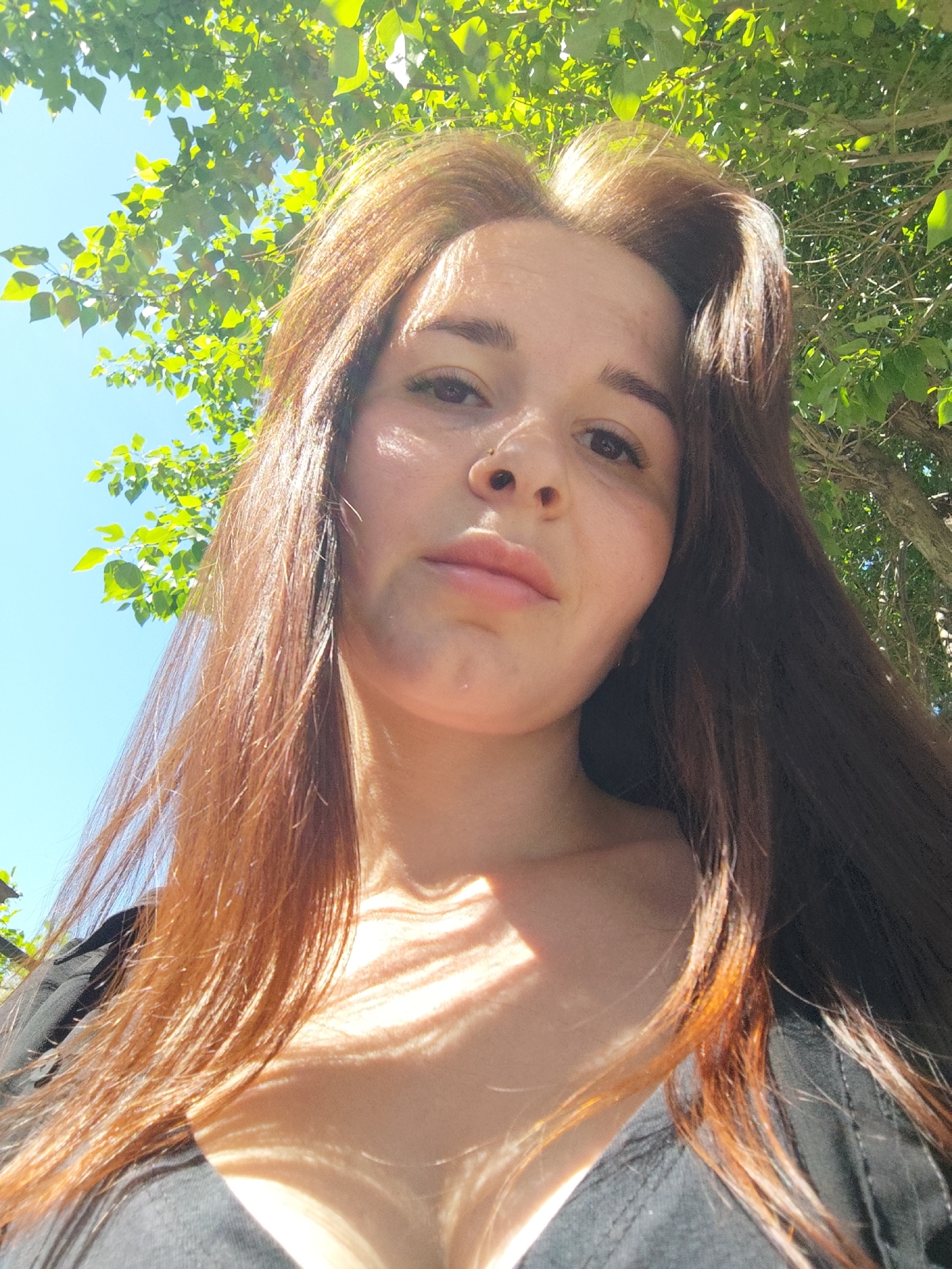 A low-angle, close-up photograph shows a woman with long brown and auburn hair standing outdoors. Above and behind her is a tree with green leaves against a bright blue sky. Light filters through the leaves, creating dappled shadows on her face, neck, and chest. She has a nose ring. She wears a black top. The location is the city of Chaikovsky, Russia.