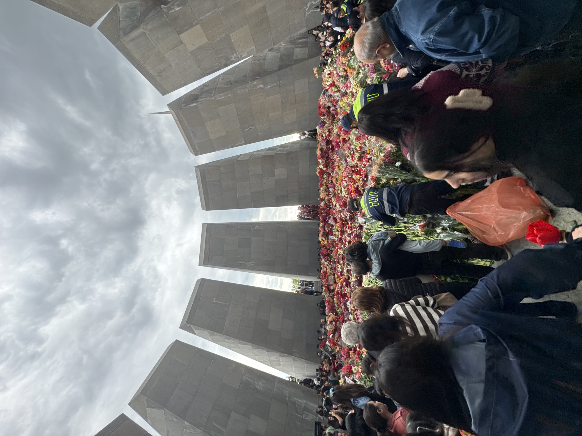 The image depicts a large, solemn gathering of people at a monumental outdoor memorial, identified by the provided context as likely being in Yerevan, Armenia. The scene is set under an overcast, cloudy sky.
The primary focus is on a vast crowd of individuals, many of whom are seen bending over to place flowers. These flowers, primarily red, pink, yellow, and white, form an extensive, dense carpet covering a significant area of the ground, indicating a collective act of remembrance or tribute. People of various ages are present, dressed in casual attire. Some individuals wear dark blue jackets or vests with partially visible white numbers "112" or "102" and Armenian script, possibly indicating official personnel or event staff. An orange plastic bag is also visible among the flowers.
In the background, several tall, imposing grey stone steles or slabs rise upwards. These angular, geometric structures are characteristic of a memorial monument, and their arrangement suggests a circular or semi-circular formation. A slender, pointed spire or monument element can be faintly discerned against the cloudy sky between some of these steles.
The activity taking place is a large-scale floral tribute, common at memorial sites, suggesting an event of national or historical significance. The overcast weather contributes to a somber and respectful atmosphere. The architecture, the sheer volume of flowers, and the respectful posture of the crowd all convey a strong sense of reverence and collective memory.