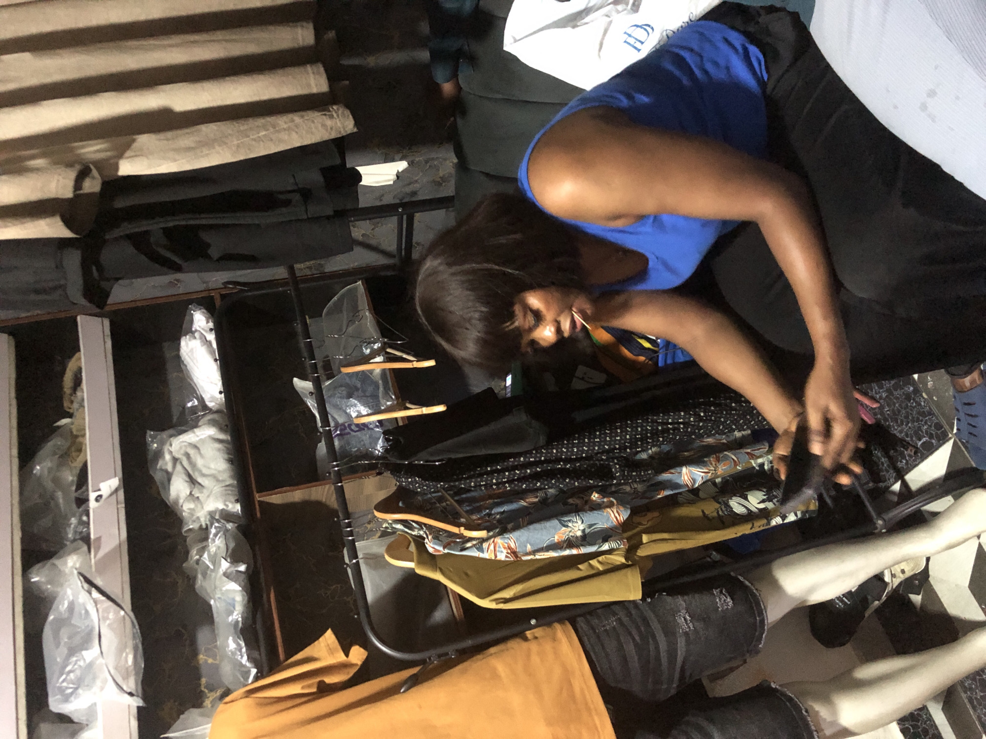 A young woman with a dark bob haircut, wearing a blue tank top and black pants, sits on a dark green couch in what appears to be a store in Ado, Nigeria. She is holding a phone in her hands and a toothpick is in her mouth. In the background, clothing racks are filled with garments, and shelves are stacked with items wrapped in plastic. To the left, a mannequin dressed in a mustard-colored t-shirt and distressed black denim shorts stands next to the woman. The overall environment suggests a retail setting, possibly a boutique or clothing store.