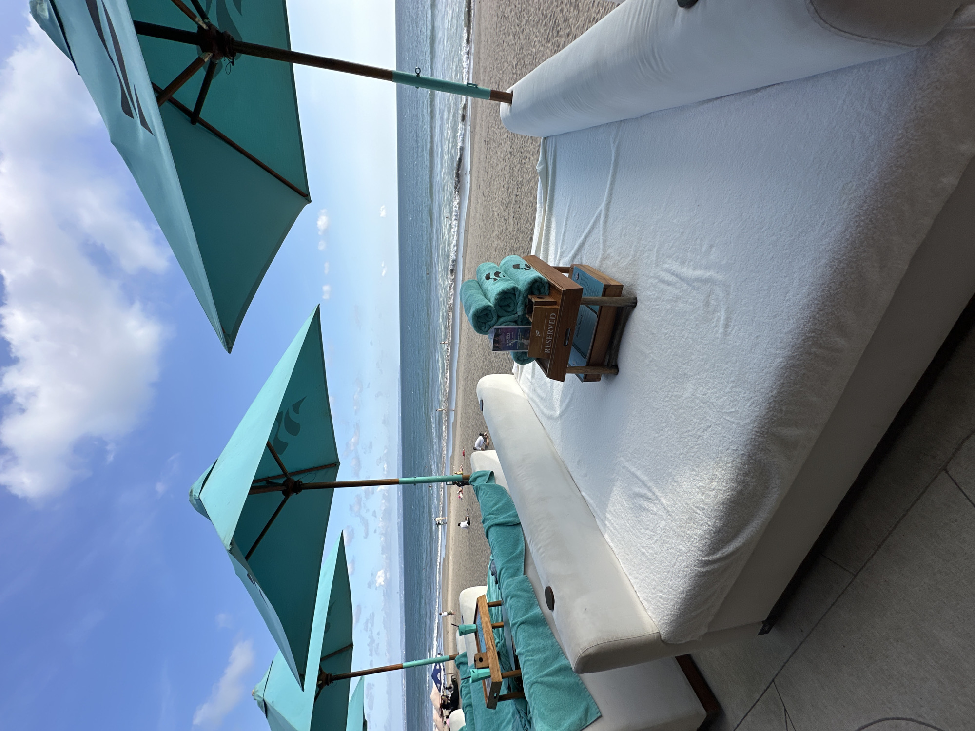 The image shows a beach scene at a resort in North Kuta, Indonesia. Several turquoise-colored umbrellas are visible in the left foreground, angled upwards toward a partly cloudy sky. A white daybed with a white mattress is placed adjacent to the umbrellas. On the daybed, a wooden tray holds rolled-up turquoise towels and informational material. In the background, there is a sandy beach with people walking along the shoreline. The ocean is visible behind the beach.
