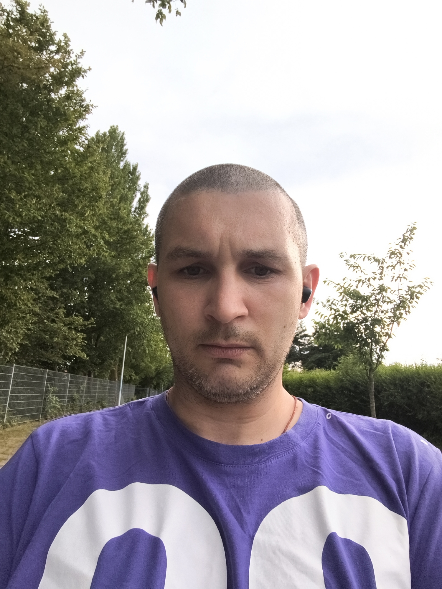 In an outdoor, daytime scene, a person is pictured from the chest up, facing the camera directly. They have short, shaved hair and appear to be wearing black wireless earbuds. Their expression is neutral, and they have a light beard. The person is wearing a purple t-shirt with a large white graphic, partially visible. In the background, there are trees, a metal fence, and a hedgerow. The sky is overcast.
The image was taken at the Marina in Wolgast, Germany, along the Peene River. The specific location is near the promenade, with the marina's yachts and boats visible in the distance to the right. The trees in the background are typical of the region's vegetation. The overall setting is that of a park or recreational area within the city.