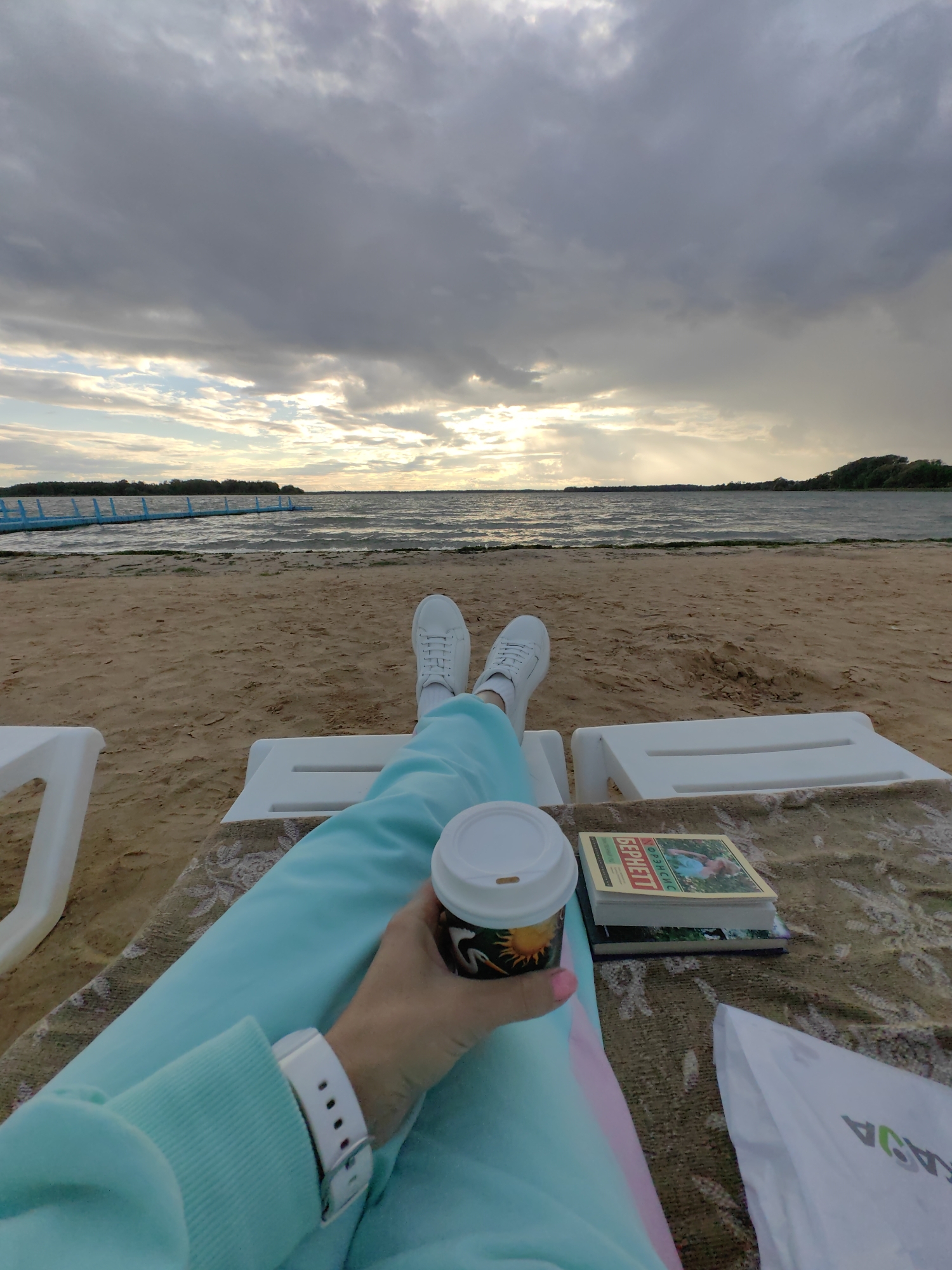 The image depicts a person relaxing on a beach in Myadzyel, Belarus. The perspective is from the viewpoint of the person, looking out at a body of water and a cloudy sky. The foreground shows the person's legs, clad in light blue sweatpants, and white sneakers. A white smartwatch is visible on the left wrist. The person is holding a paper cup with a lid, decorated with a sun graphic. Next to the cup are two books and a patterned blanket. In the mid-ground, a sandy beach stretches towards the water, with a blue pier extending into the lake. The background features the lake, a distant treeline, and a dramatic sky with heavy clouds and glimpses of sunlight. The environment suggests an overcast day at a lakeside resort.