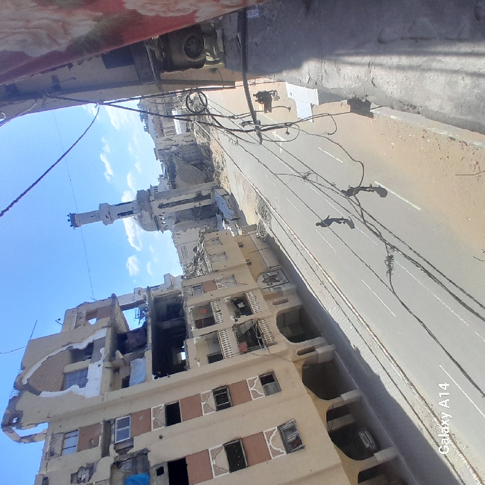 The image displays a severely damaged urban street scene in Khan Yunis, Palestinian Territory, captured from an elevated vantage point on a clear, sunny day. A multi-story building on the left exhibits extensive structural damage, with several upper floors partially collapsed, exposing interior rooms and twisted rebar. Its lower levels feature balconies and arched ground-floor entrances, with one light-colored vehicle parked beneath the arches. Further down the street, other buildings also show damage. In the distance, a mosque with a tall minaret and a dome stands amidst rubble, appearing significantly damaged. Two individuals, dressed in dark clothing, walk on the paved road, which features white lane markings, away from the viewer. They cast elongated shadows on the asphalt. The unpaved sides of the road consist of dirt and debris. Electrical or communication cables are visible traversing the frame diagonally in the foreground. A patterned fabric partially obscures the immediate right edge of the image.