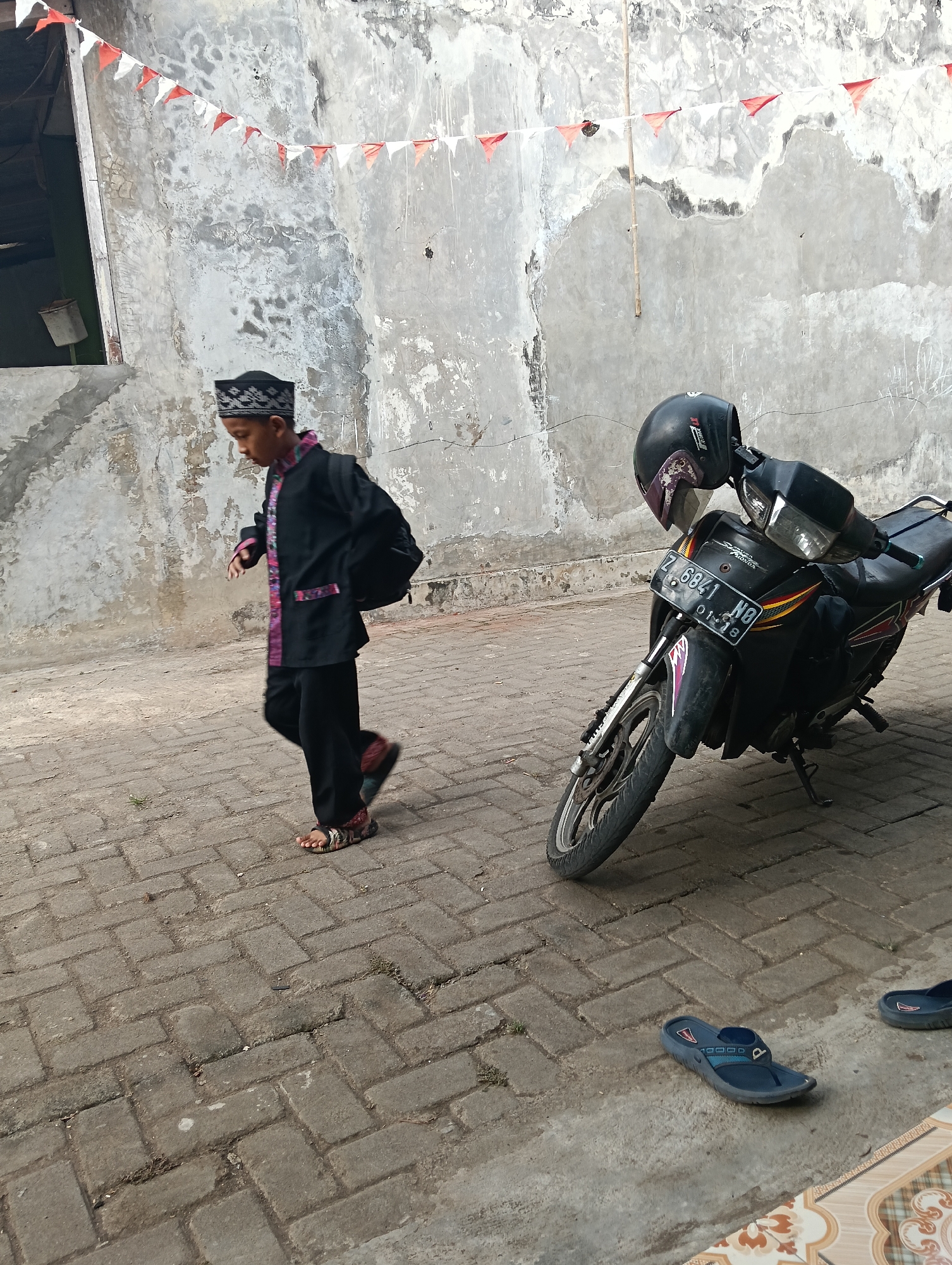 An outdoor, ground-level, medium shot captures a young boy in traditional clothing walking on a brick-paved walkway. The boy is wearing a black, long-sleeved shirt adorned with colorful patterned trim and black pants, along with a black cap. He has a black backpack slung over his right shoulder. To the right of the boy is a black motorcycle with a black helmet placed on its handlebars. The motorcycle has a license plate that reads "Z 6841 NO" and a date sticker showing "01-18". In the background, a textured, light-colored wall is visible, decorated with a string of small red and white triangular flags. The ground is paved with interlocking grey bricks. In the lower right corner, a pair of dark blue flip-flops lie on the ground. This scene is consistent with the general appearance of an outdoor area in a residential or public space within Tasikmalaya, Indonesia, potentially decorated for a local celebration.