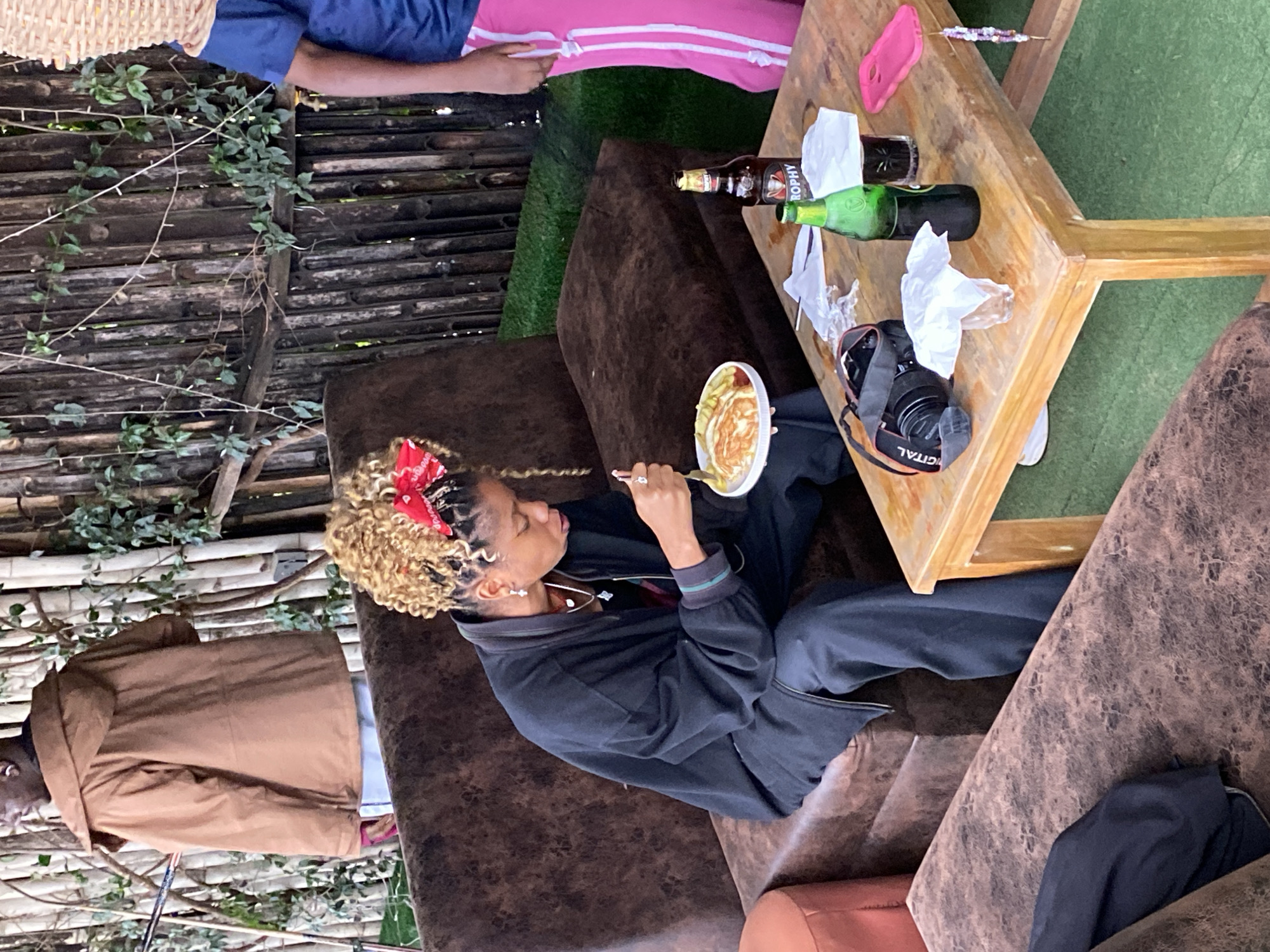 A medium shot, captured at an outdoor, informal setting in Gura Top, Nigeria, shows a young Black woman with blonde, braided hair, wearing a red bandana, black jacket, and dark pants, seated on a brown couch. She is eating from a white plate of food with a fork, her gaze focused downwards. On a wooden table in front of her, there's a camera, a green bottle, a brown bottle with a red label reading "TROPHY," a clear glass with dark liquid, and napkins. A person in a brown jacket stands in the background, partially obscured by bamboo stalks. The environment includes green grass and a wooden fence.