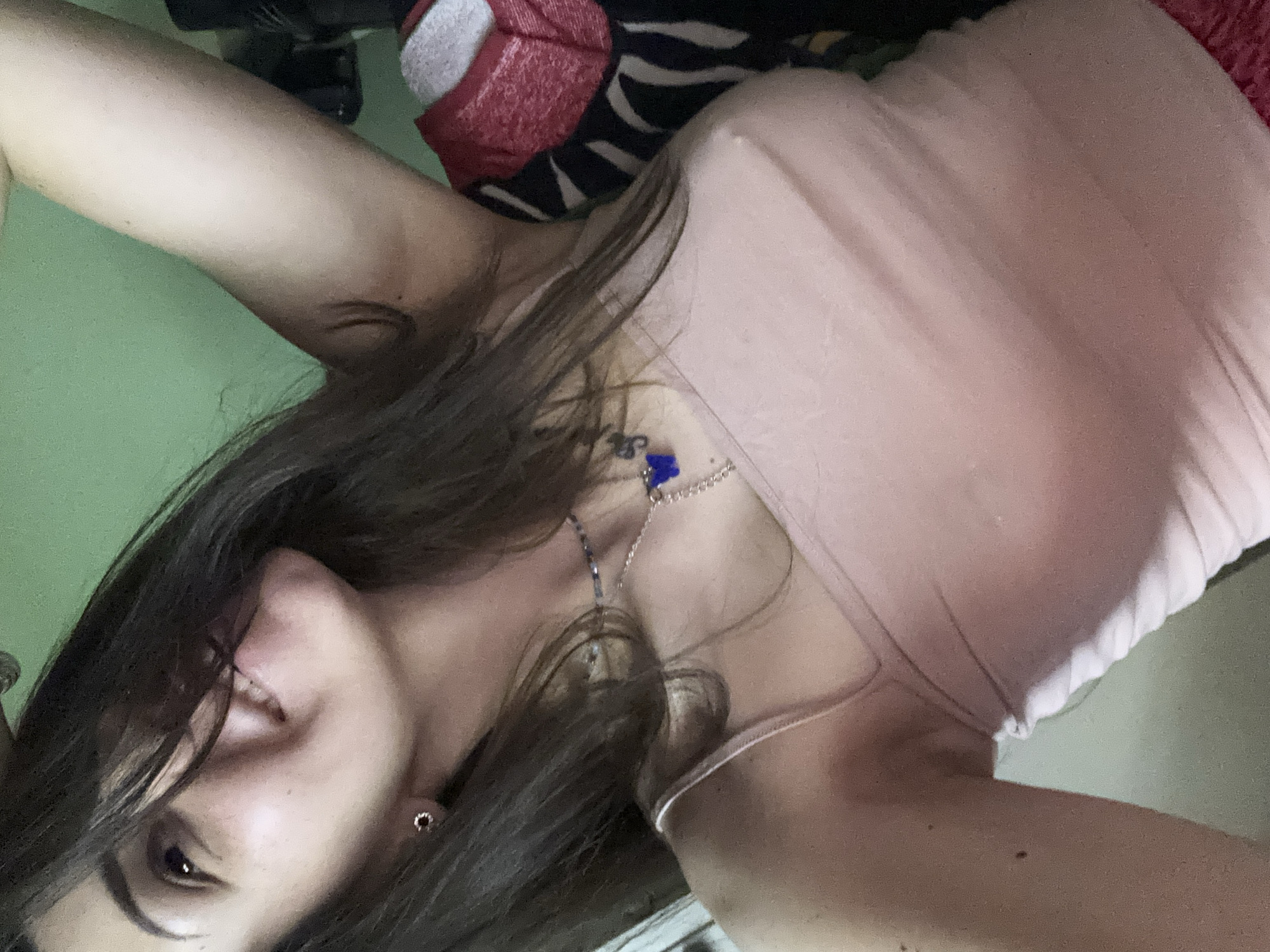 A young woman is captured in a close-up, diagonal selfie, set against an indoor backdrop in Sofia, Bulgaria. She has long, dark brown hair that frames her face and shoulders. Her head is tilted, and she looks towards the camera with a gentle smile, revealing a hint of teeth. She wears a light pink or nude-colored tank top with thin straps and a delicate silver-toned necklace featuring a small blue charm, possibly an evil eye. A dark stud earring is visible in her right ear.
Her left arm is raised above her head, mostly out of frame, suggesting a relaxed pose, possibly lying down. The background on the left features a plain, light green wall. On the right, a mix of patterned fabrics, including black and white stripes (reminiscent of zebra print) and a section of textured red material, likely part of bedding or furniture, fills the space. The lighting is soft and indoor, creating subtle shadows. The overall mood conveyed is calm and relaxed. There is no visible text in the image.