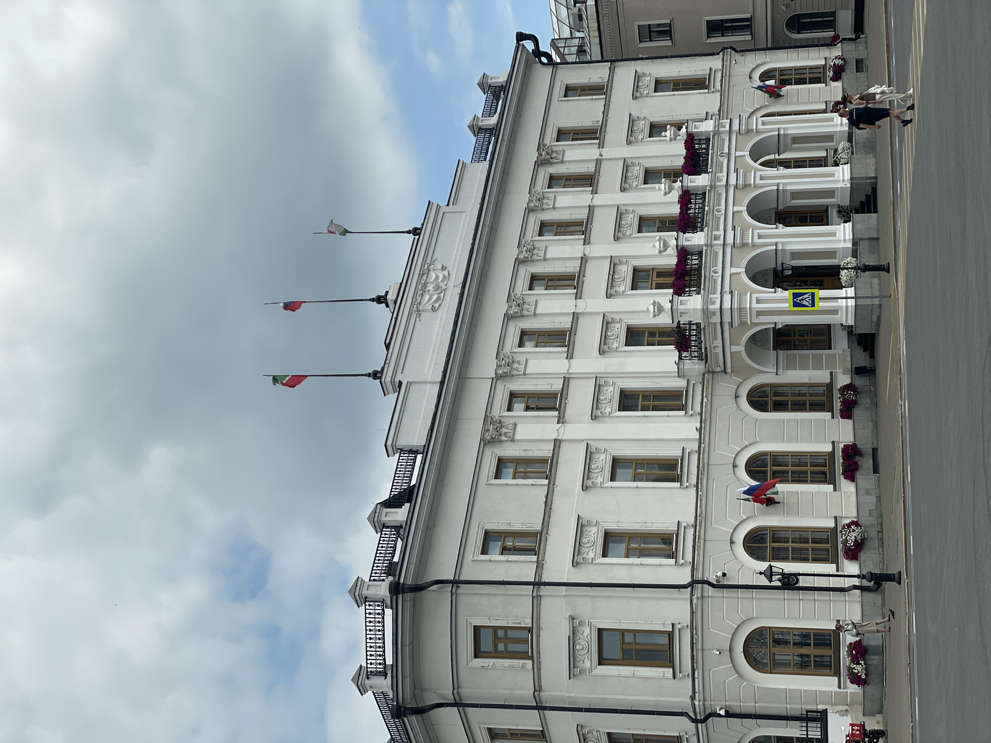 The image shows the National Museum of the Republic of Tatarstan in Kazan, Russia. The building is a grand, white, three-story structure with ornate architectural details, including columns, decorative moldings, and a balustrade along the roofline. Several flags are visible, including the Russian flag and what appears to be the flag of Tatarstan. The facade features numerous arched windows. In front of the building, there is a paved area with a crosswalk where two individuals are walking. Flower planters adorn the building's lower level, and a lamppost stands on the sidewalk. The sky is partly cloudy.
