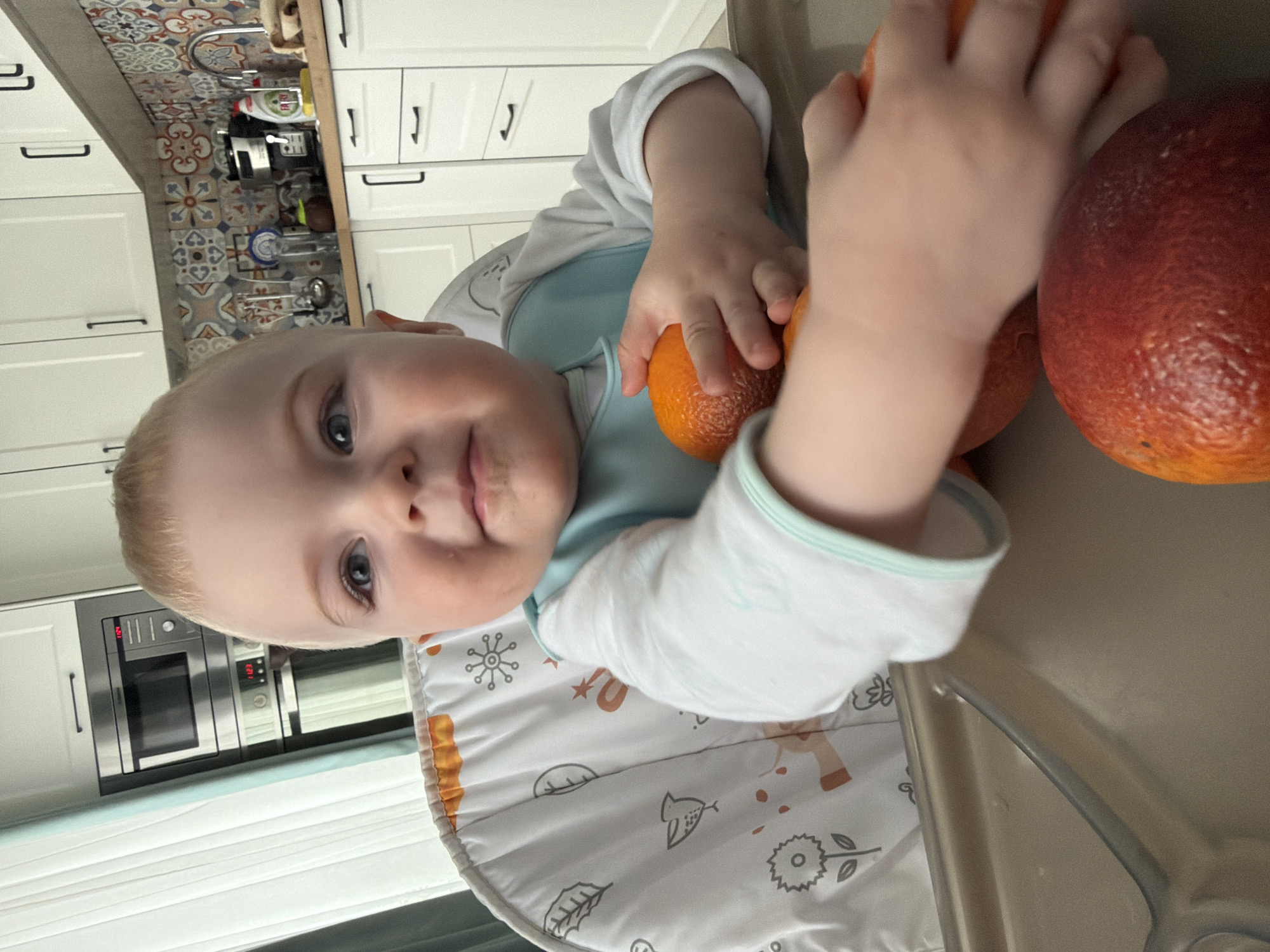 A baby with blond hair and blue eyes sits in a high chair with a bib. The baby is touching two oranges. The scene is indoors, in a kitchen with white cabinets, a microwave, and patterned tiles on the backsplash. The location is not discernible as being specifically in Votkinsk, Russia.