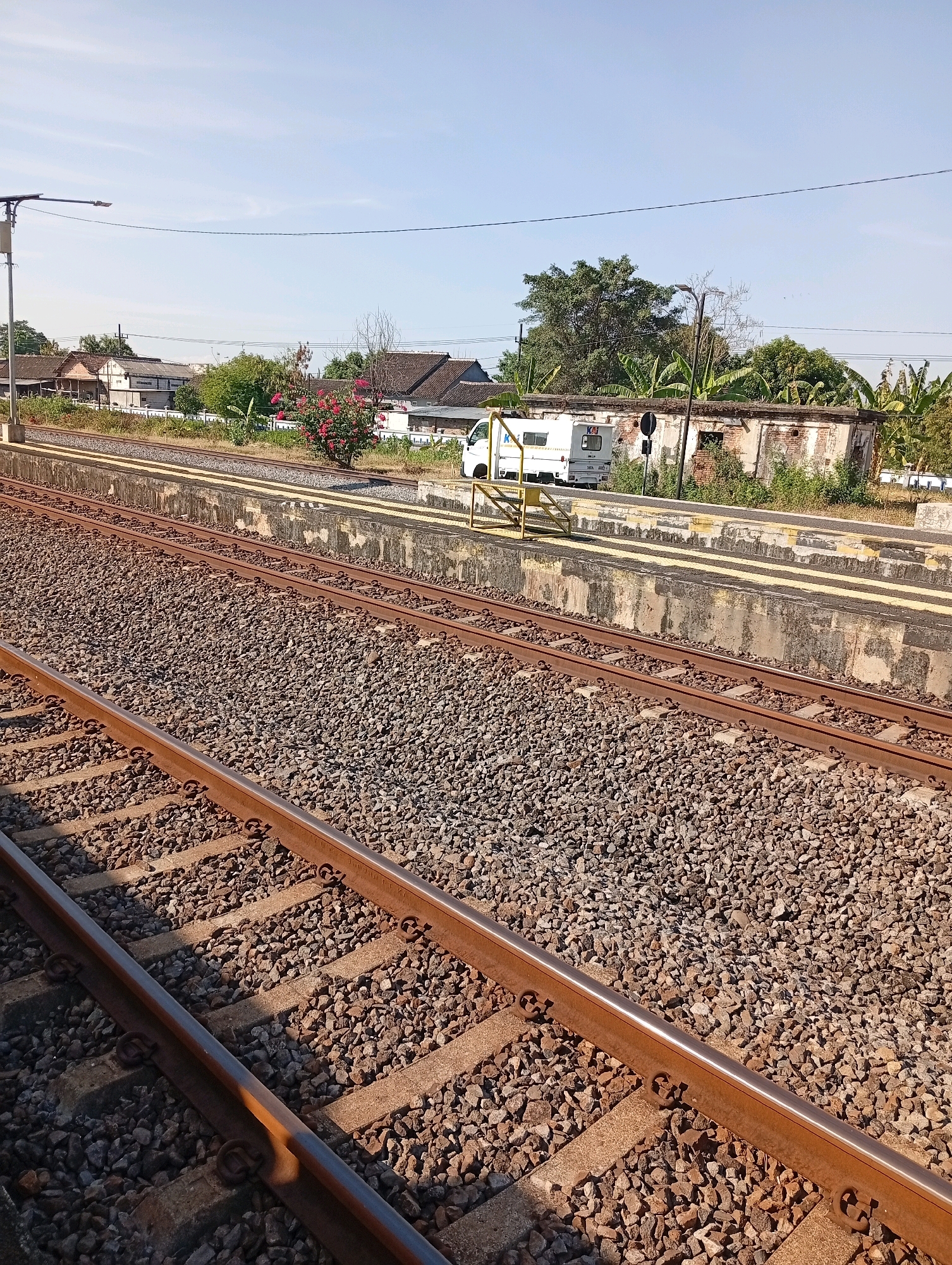 The image depicts a railway station in Tebon, Indonesia. In the foreground, there are two sets of train tracks composed of weathered metal rails resting on concrete sleepers and covered with gravel. A concrete platform runs parallel to the tracks, marked with yellow lines. Beyond the platform, a white utility vehicle with the word "KA" (likely for Kereta Api, Indonesian for train) is parked. Behind the vehicle, an old, partially dilapidated brick building stands, with vegetation growing around it. In the background, several buildings with tiled roofs, trees, and utility poles are visible under a clear blue sky. The scene suggests a sunny day at a rural or suburban train station.