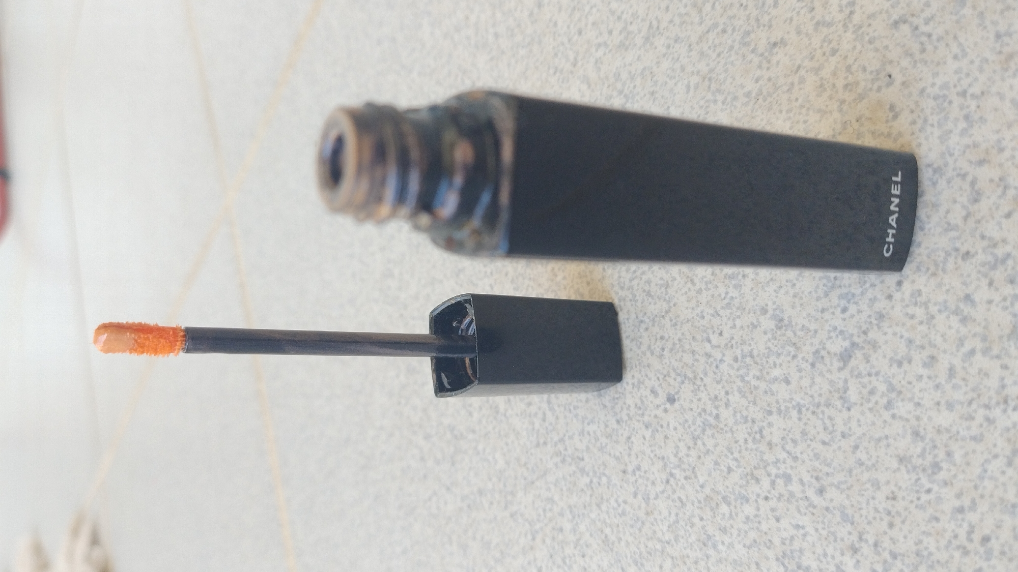The image shows a close-up, eye-level shot of an open matte black lipstick tube with its applicator wand beside it. The applicator wand, which has a fuzzy orange tip with a small amount of light peach lipstick on it, is extended out of its cap, which is placed on a light-colored, speckled tiled surface. The lipstick tube itself is dark and appears to have some product residue around the rim. The word "CHANEL" is visible in white text on the bottom of the tube. The background is out of focus, showing more of the speckled tiled surface. The scene is indoors, possibly on a floor. There are no people or animals in the image. The image provides no information about the location in the city of Perea, Greece.