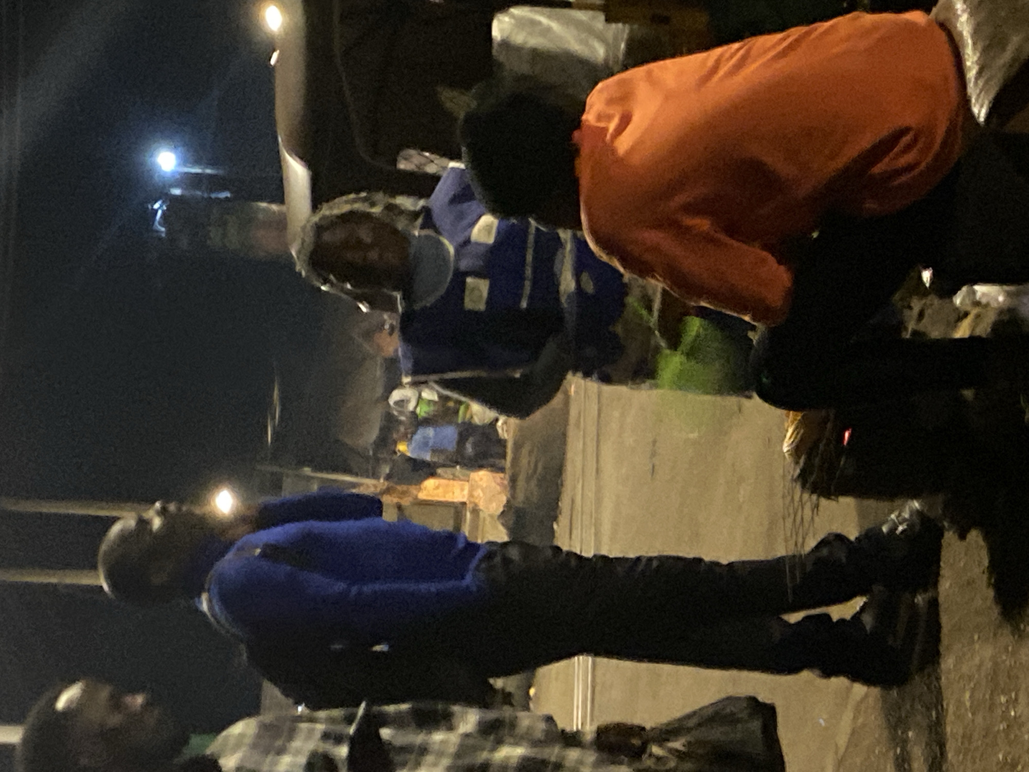 This media file captures a nighttime scene, likely a street or market setting in Bukuru, Nigeria, illuminated by artificial lights. Several individuals are visible, engaged in various activities.
In the mid-ground, a person wearing a blue jacket or long-sleeved top and dark trousers stands upright, facing right. Their bald head is visible, and they appear to be looking towards something or someone out of frame. Another individual, partially visible in the foreground to the lower left, also appears to be standing or seated.
Towards the right side of the frame, another person is prominently featured, clad in a bright orange long-sleeved shirt and dark trousers. This person is leaning or bending forward significantly, suggesting interaction with an object or activity at a lower level. Behind them, a person with dreadlocks or braids is wearing a dark blue vest with white patches or logos, possibly a uniform. This individual is looking towards the person in orange or the activity they are engaged in.
The background is dark but shows glimmers of distant bright lights, possibly streetlights or vehicle lights, confirming the nighttime setting. There are also indistinct forms and structures, suggesting market stalls, buildings, or clutter common in urban environments. The ground appears to be concrete or packed earth.
The overall impression is one of a bustling nocturnal environment where people are gathered, possibly working or socializing. No specific text is clearly visible.