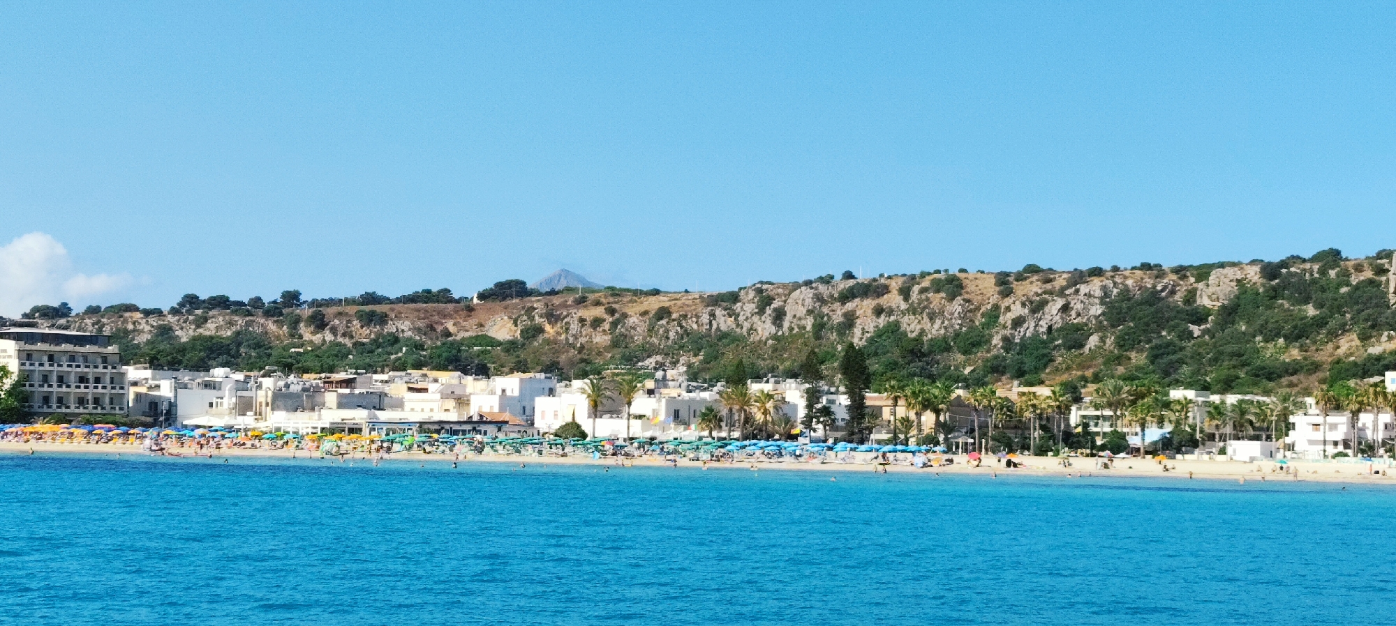 This is a wide, eye-level, horizontal photograph capturing the coastline of San Vito Lo Capo, Italy. The foreground is dominated by vibrant, turquoise water. The middle ground features a sandy beach filled with numerous colorful umbrellas and beachgoers enjoying a sunny day. Behind the beach, a cluster of white buildings, typical of Mediterranean architecture, are visible. Further in the background, a lush green hillside rises, dotted with rocky outcrops and trees. The sky above is a clear, bright blue. In the distance, a hazy mountain peak can be seen. The scene conveys a lively and inviting atmosphere characteristic of a popular beach resort.