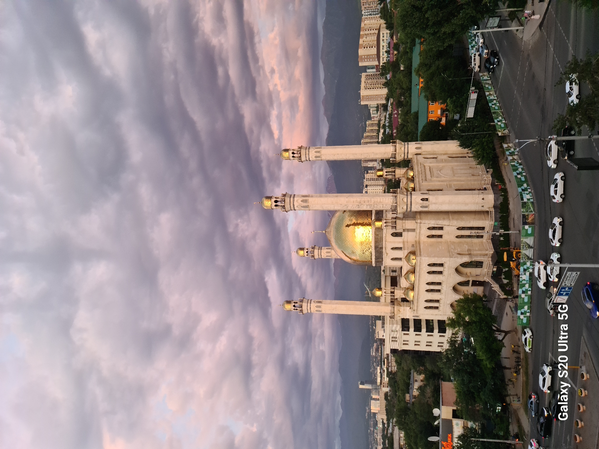 This is a vertical, high-angle photograph of the Central State Mosque in Almaty, Kazakhstan. The mosque, with its white facade and golden domes and minarets, is the primary subject, illuminated by the warm light of what appears to be sunset or sunrise. The sky is filled with dramatic clouds, streaked with hues of purple, pink, and grey, suggesting dynamic weather conditions.
In the background, a cityscape is visible, with numerous apartment buildings and some trees. The architecture suggests a modern urban environment. A street runs alongside the mosque, with several cars driving and parked, indicating that the mosque is accessible by road and is likely a functional religious site. The presence of traffic suggests a normal day in the city.
Notable details include the intricate design of the mosque's minarets and the shimmering golden dome, which catches the light. The cloudy sky adds a sense of drama and beauty to the scene, evoking a contemplative or serene emotion. The overall impression is one of grandeur and tranquility amidst an urban setting.
The timestamp "Galaxy S20 Ultra 5G" is visible in the lower right corner of the image, indicating the device used for capture. There is also some unreadable text on a sign in the distance. The time of day is likely late afternoon or early evening, given the lighting and cloud colors.