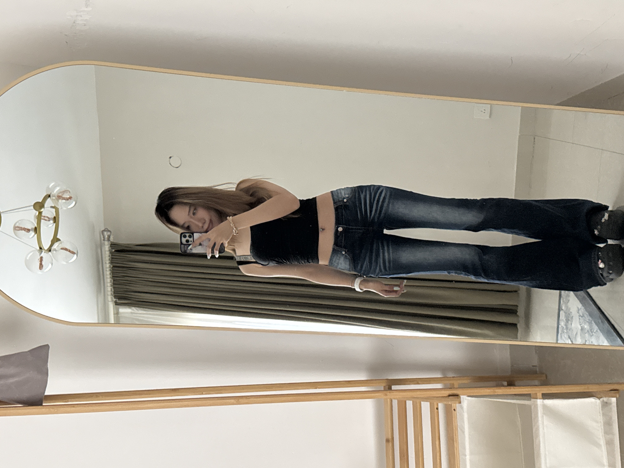 In Yuzhong District, China, a young East Asian woman stands in front of a full-length mirror, taking a selfie. She is wearing a black cropped tank top, dark wash flared jeans, and black platform Crocs. A silver belly button ring is visible. She is accessorized with a white beaded bracelet on her left wrist and a beaded bracelet on her right wrist. Her hair is long and brown, and she is looking at her phone. The mirror reflects a curtained window and a wall with a light switch. To the left of the mirror, a wooden garment rack with white fabric bins is visible. The room has tiled flooring and the lighting appears to be natural, coming from an unseen source.
