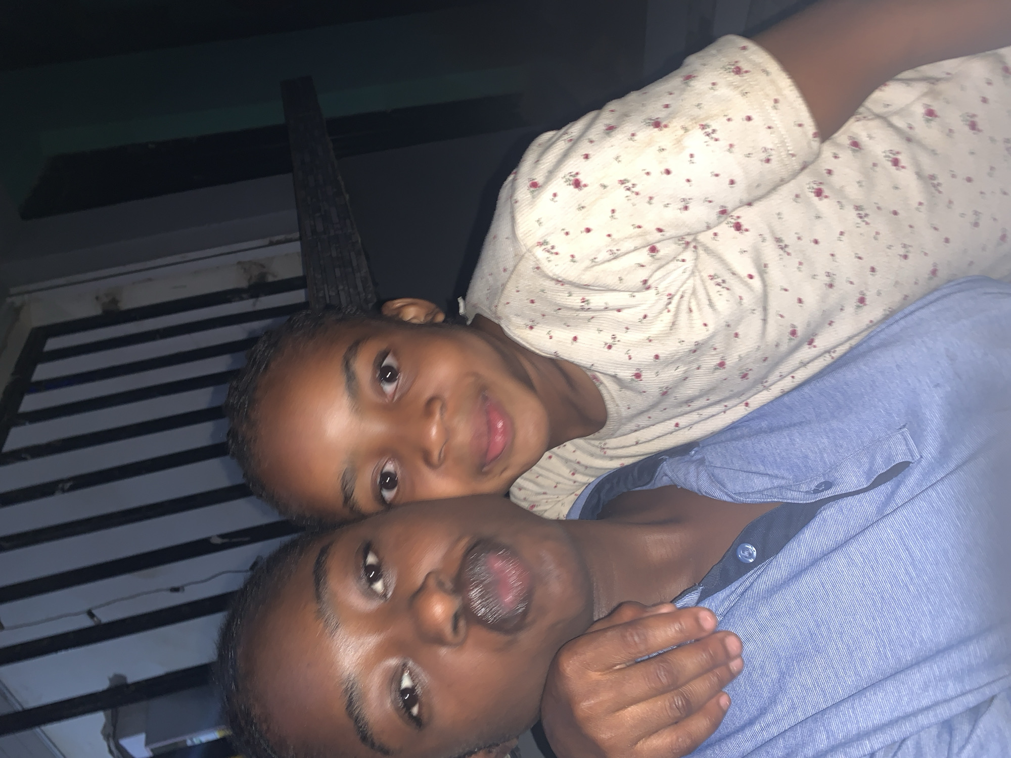 Two individuals, one wearing a white shirt with small red floral patterns and the other wearing a blue button-down shirt, are positioned closely together. The individual in the white shirt is slightly behind the individual in the blue shirt. The setting appears to be outdoors at night, based on the low lighting and the presence of a dark structure in the background, potentially a building or fence, with horizontal slats. The photo was taken in New Stadium, Nigeria.