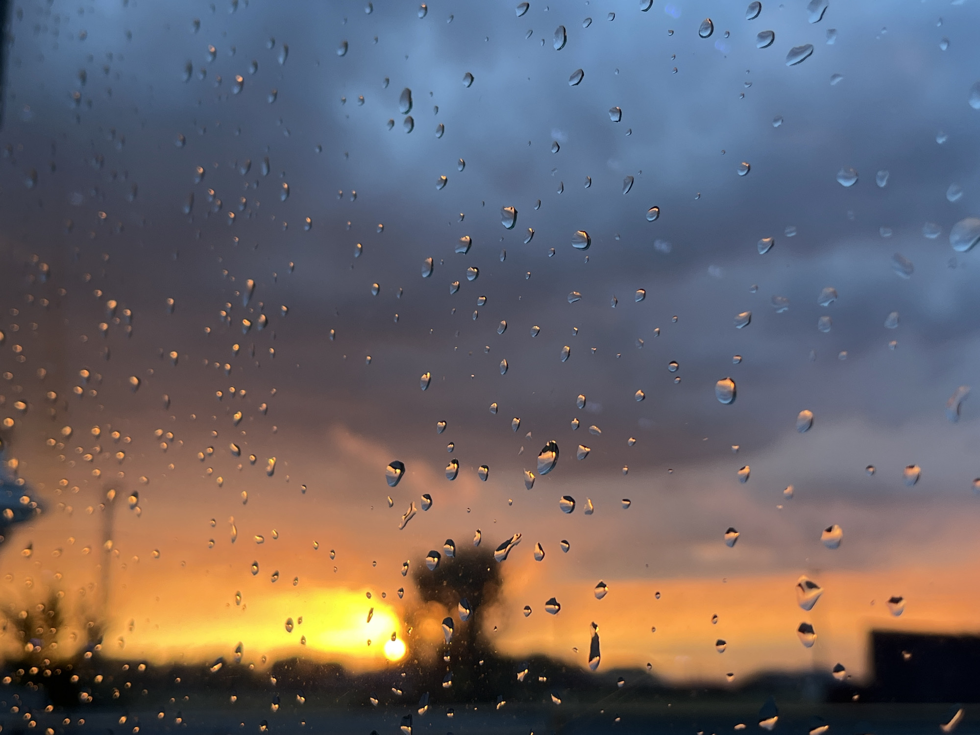 The image captures a view through a rain-streaked window during a sunset. The foreground is dominated by numerous water droplets on the glass, which distort the view of the background. Through the droplets, a vibrant sunset can be seen, with hues of orange and yellow illuminating the sky. The sun itself is partially obscured, casting a warm glow. In the distance, silhouetted against the sunset, is a dark, bare tree, and a dimly lit outline of what appears to be a building or structure. The sky above the sunset is a gradient of darker blue and grey, with cloudy textures visible. The lower portion of the image shows a darker, out-of-focus strip, likely representing the ground or a ledge. No people or specific identifiable landmarks of Taganrog City District, Russia, are discernible in the image; it is a generalized scene of a sunset viewed through a wet window.