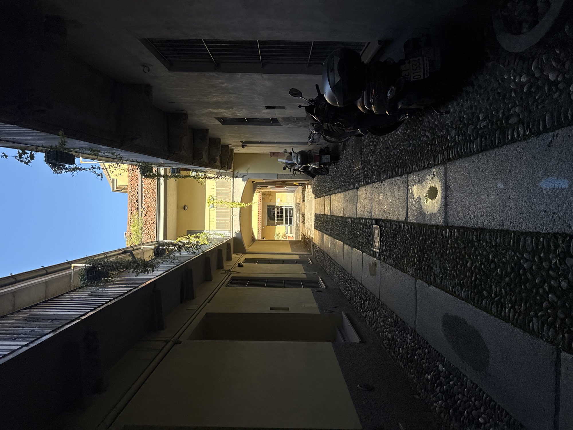 A narrow, high-walled alleyway or courtyard is depicted, looking into the distance. The scene is characterized by strong contrasts between deep shadows and bright sunlight, suggesting a clear day, likely morning or late afternoon.
The ground of the alley features a distinctive pattern of small, dark pebbles, similar to cobblestones, separated by smooth, light-colored stone strips. Patches of sunlight illuminate various sections of this patterned ground. Along the right side of the alley, several scooters or motorcycles are parked. The license plate of one scooter is partially visible, showing "E1 01534".
Tall buildings line both sides of the alley, creating a canyon-like effect, with many sections bathed in shadow. The building walls appear to be made of stone or concrete, with some areas painted in light yellow or beige tones. Multiple windows, some fitted with security bars or grates, are visible. On the left side, plants and vines grow along railings and cascade from upper levels. A clear blue sky is visible at the top left of the frame. At the far end of the alley, an archway or opening leads to a brightly lit area. The architectural style and presence of scooters are consistent with the provided location context of Milan, Italy.