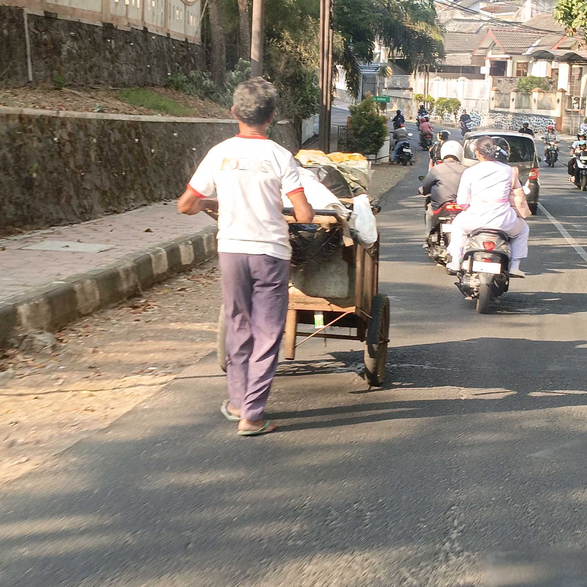 A person with gray hair, wearing a white t-shirt with red trim and purple pants, pushes a two-wheeled cart filled with objects. The cart has a wooden frame and appears to contain bags and fabric. The person is walking on a paved road, and the cart's wheels are visible. In the background, there is a retaining wall made of rough stones, a sidewalk, and trees. Further in the distance, there are buildings with tiled roofs and other people on motorcycles. This scene is consistent with a street in the city of Salatiga, Indonesia.