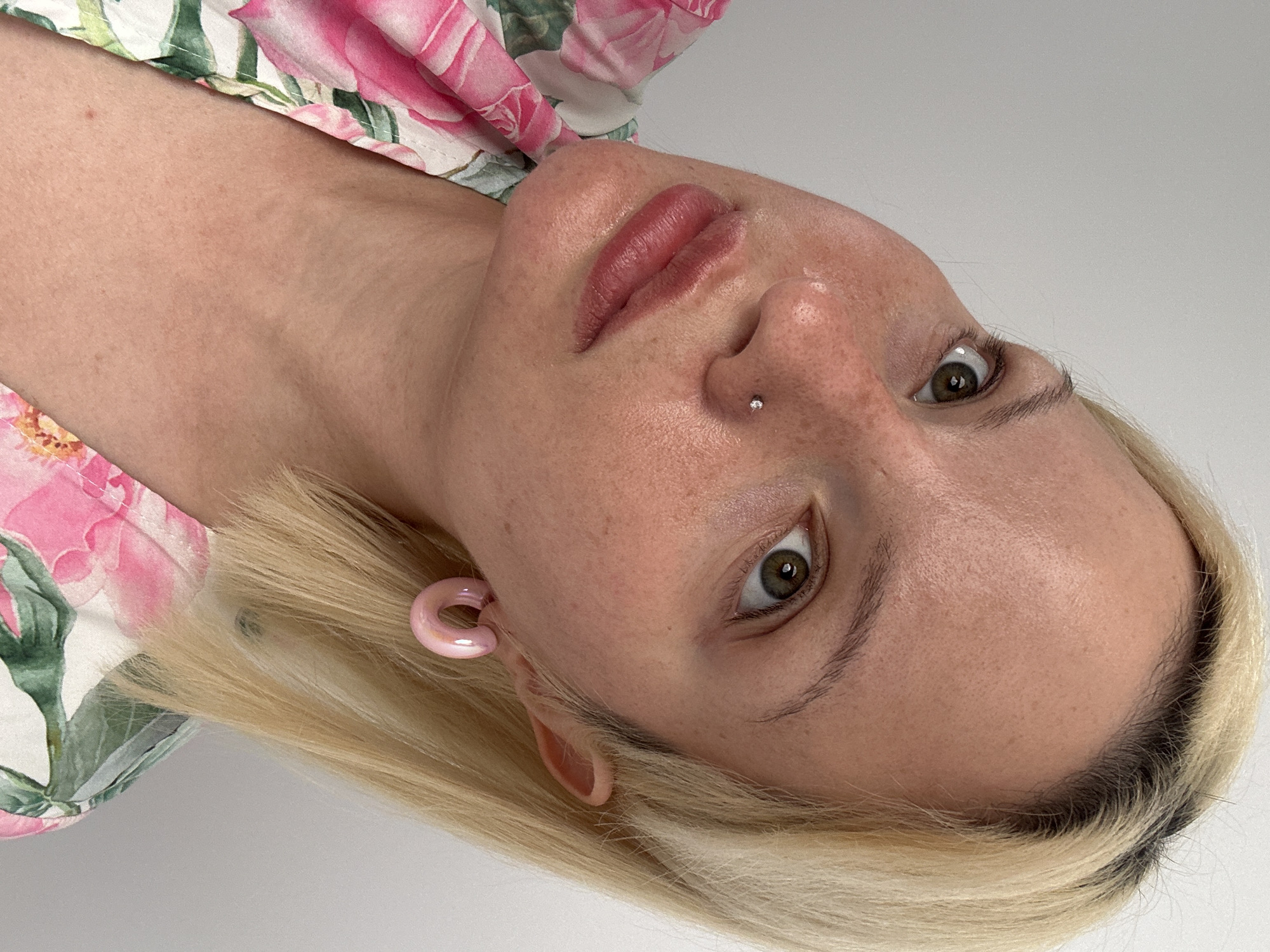 This close-up portrait features a person, likely female, with fair skin and short blonde hair, presented in an indoor setting with a plain, light-colored background. The image is rotated, showing the subject's head tilted, with her eyes looking directly at the camera.
The subject has light brown or green eyes and full lips with a subtle pink or berry-toned lip color. A small, clear stud piercing is visible on her nose. Her skin shows natural texture, including visible pores and faint freckles or sunspots, with a slight sheen on the forehead. Her blonde hair is short, possibly a bob, with darker roots visible near the scalp.
She wears a light pink, C-shaped earring in one ear, appearing to be made of a smooth material like ceramic or plastic. Part of a garment with a vibrant pink floral pattern, featuring roses and green leaves, is visible on her shoulder and chest. The overall impression is a detailed, natural portrait, focusing on facial features, skin texture, and subtle accessories.
No specific activity is depicted beyond posing for the photograph. The plain indoor background provides no discernible location cues to Budva, Montenegro, nor any indication of the time of day or specific weather conditions. There is no visible text in the image.