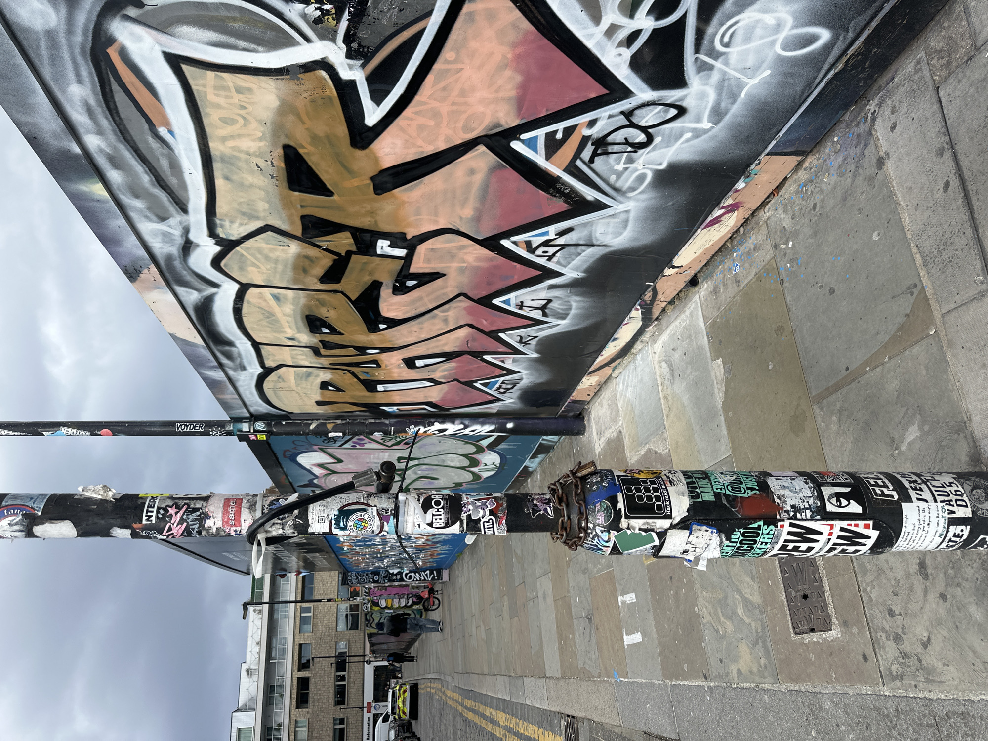 The image displays an urban street scene in London, United Kingdom, under an overcast grey sky. In the foreground, a prominent black pole is densely covered with numerous diverse stickers and some graffiti tags. A rusted metal chain with a padlock is wrapped around its lower section, and a black cable is secured to its upper portion by a white plastic clip. A second, thinner black pole, also adorned with stickers, stands further back to the right.
To the right of the poles, a tall wall extends laterally across the frame, completely covered in layered graffiti. The central artwork features large, stylized letters rendered in black, orange, brown, and red with distinct white outlines. Smaller tags and paint splatters are visible beneath and surrounding this primary graffiti, which is applied over a dark background. The lower edge of the wall reveals sections of painted wooden boarding.
The ground comprises a paved stone sidewalk with square slabs, exhibiting signs of wear, cracks, and scattered blue and white paint splatters. To the left of the sidewalk, an asphalt road features double yellow lines painted along the curb. In the background, multiple multi-story buildings, constructed from brick and lighter materials, are visible, along with a streetlamp. Several individuals are present: one person in a dark jacket and jeans walks away from the camera on the sidewalk, near a small red bicycle. Further down the street, another individual stands adjacent to a yellow and black vehicle. The scene depicts a dynamic urban environment characterized by extensive street art and pedestrian activity.