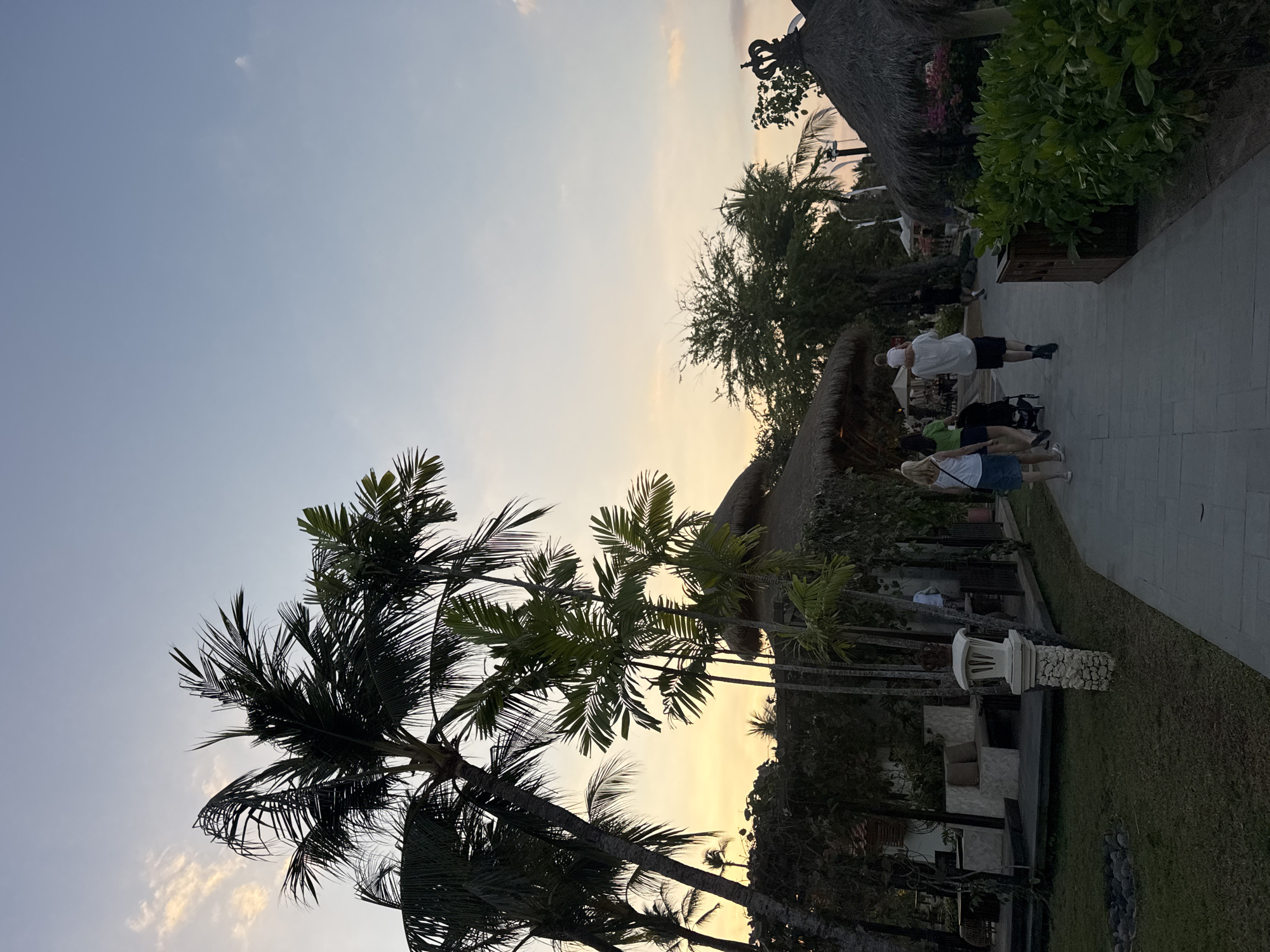 In Kuta Selatan, Indonesia, at dusk, a paved pathway winds through lush greenery and resort-like structures. Several people are visible, including a couple pushing a stroller, a man carrying a child on his shoulders, and a woman walking ahead of them. Palm trees and thatched-roof buildings dot the landscape, creating a tropical atmosphere. The sky is a soft blend of orange, yellow, and blue, indicating the setting sun. The scene captures a serene evening ambiance.