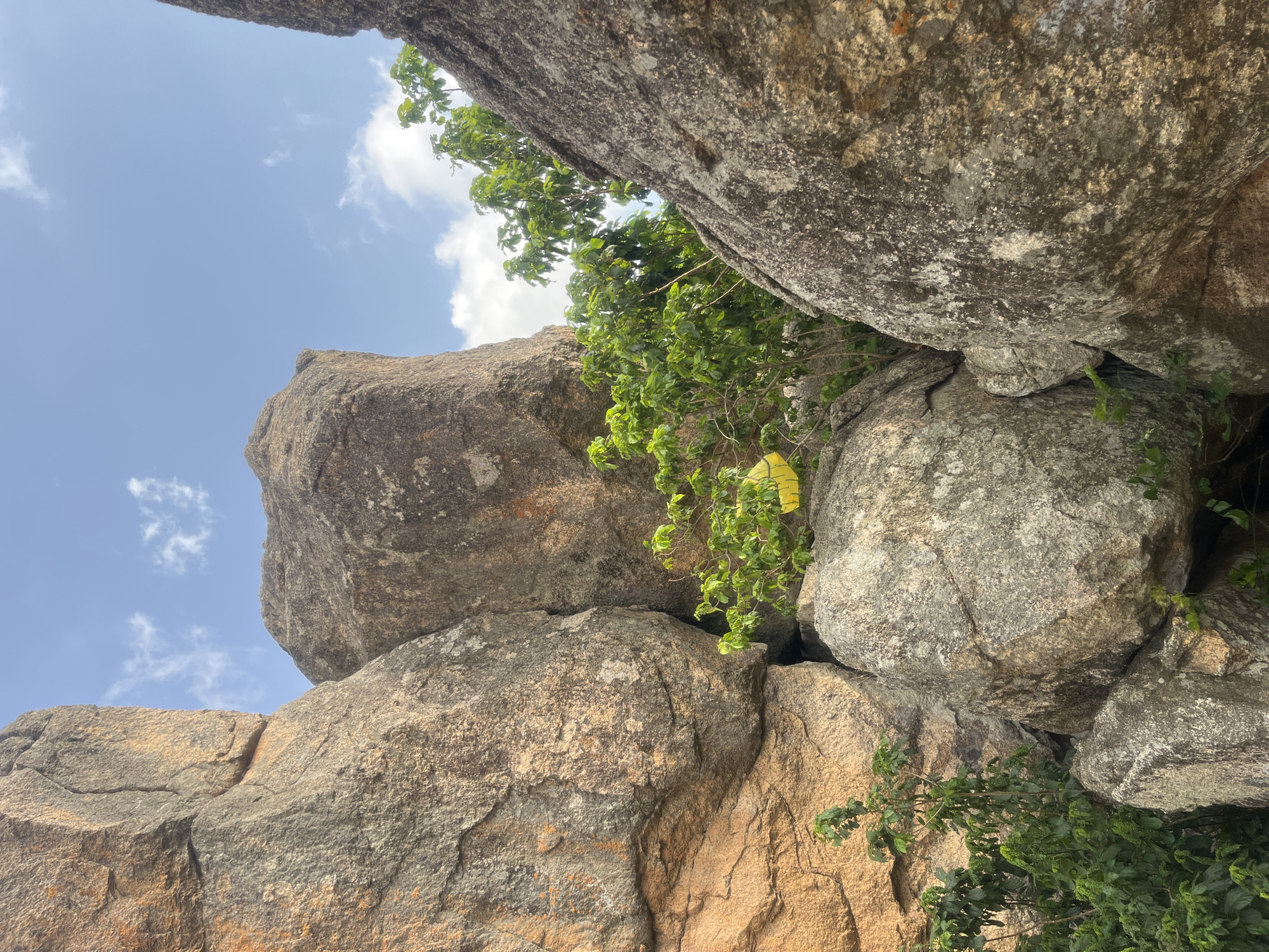 The image depicts a view upwards through a rock formation in Jos, Nigeria, known for its granite outcrops. The scene is dominated by large, stacked boulders, ranging in color from tan to gray, with patches of green vegetation sprouting between them. The sky is visible in the upper portion of the frame, showing a clear blue with scattered white clouds. The perspective suggests the photograph was taken from a low angle, looking upwards through the rock structure. There are no people or animals visible in the frame.