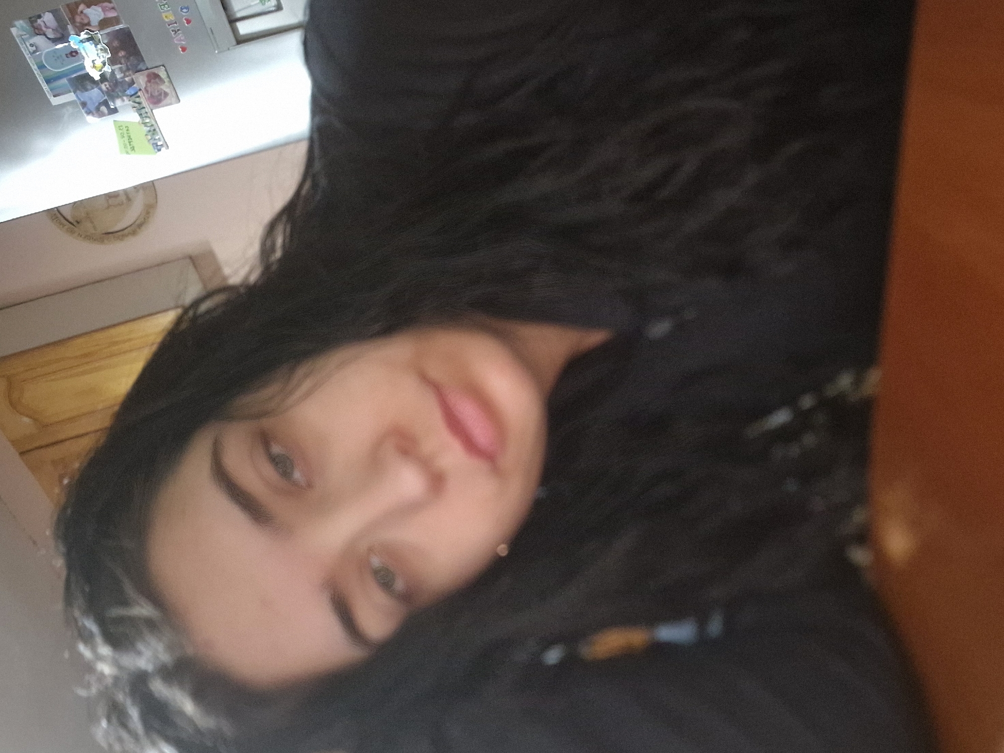 The image captures a close-up of a young woman with long, dark, wavy hair, reclining and looking directly at the camera. Her head is tilted at an angle, suggesting a selfie taken from above. She has light-colored eyes and her lips are softly parted in a neutral expression. She is wearing a dark-colored top.
The background on the left features a white refrigerator covered with several photographs, magnets, and notes. A "BIRTHDAY" decoration is visible, along with other smaller papers, including a green card with some partially legible numbers like "03--" and other indistinct text. Below the fridge, a light-colored wall and wooden details, possibly a door frame or cabinet, are present. To the right of the woman, the background is a dark, blurry area that transitions into a vertical reddish-brown surface, likely a wooden wall or furniture.
The lighting is soft and consistent with an indoor setting. The image has a slightly grainy quality, and the background elements are somewhat out of focus, emphasizing the woman's face. The overall scene conveys a casual and personal atmosphere, likely within a home. No specific activity beyond posing for a photo is taking place.