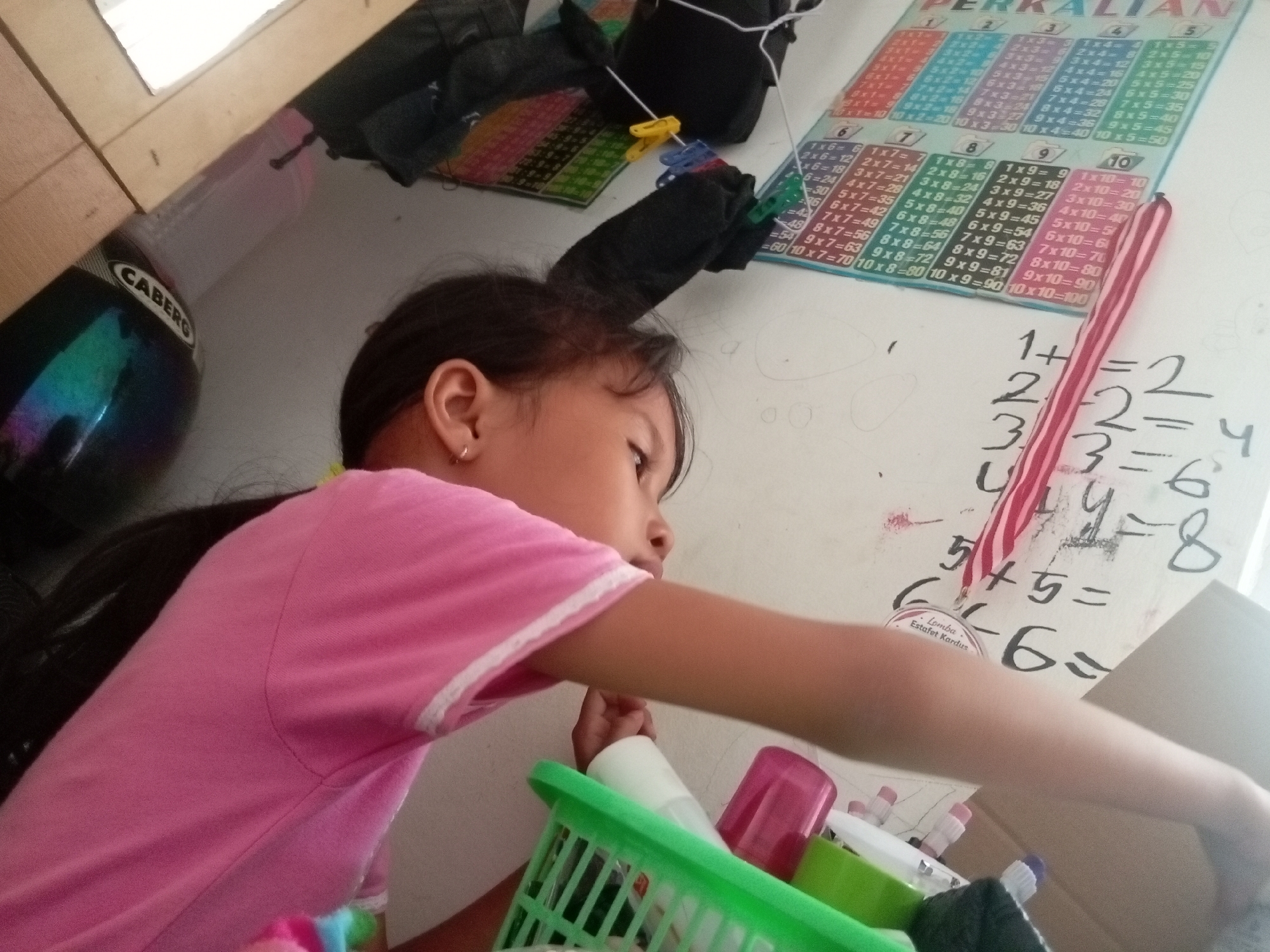 The image shows a young girl in a pink shirt looking at a whiteboard with handwritten math problems. She is sitting on the floor in what appears to be a room, possibly a study area, as indicated by the presence of educational materials like multiplication charts. A helmet with the brand "CABERG" is visible in the background, suggesting it might be an item belonging to someone in the household. The overall setting is informal and appears to be within a residential environment in Batam, Indonesia. The girl's gaze is directed downwards, and her right arm is extended, suggesting she might be interacting with something on the floor or reaching for an object. The multiplication chart on the wall displays "PERKALIAN" which is Indonesian for multiplication.