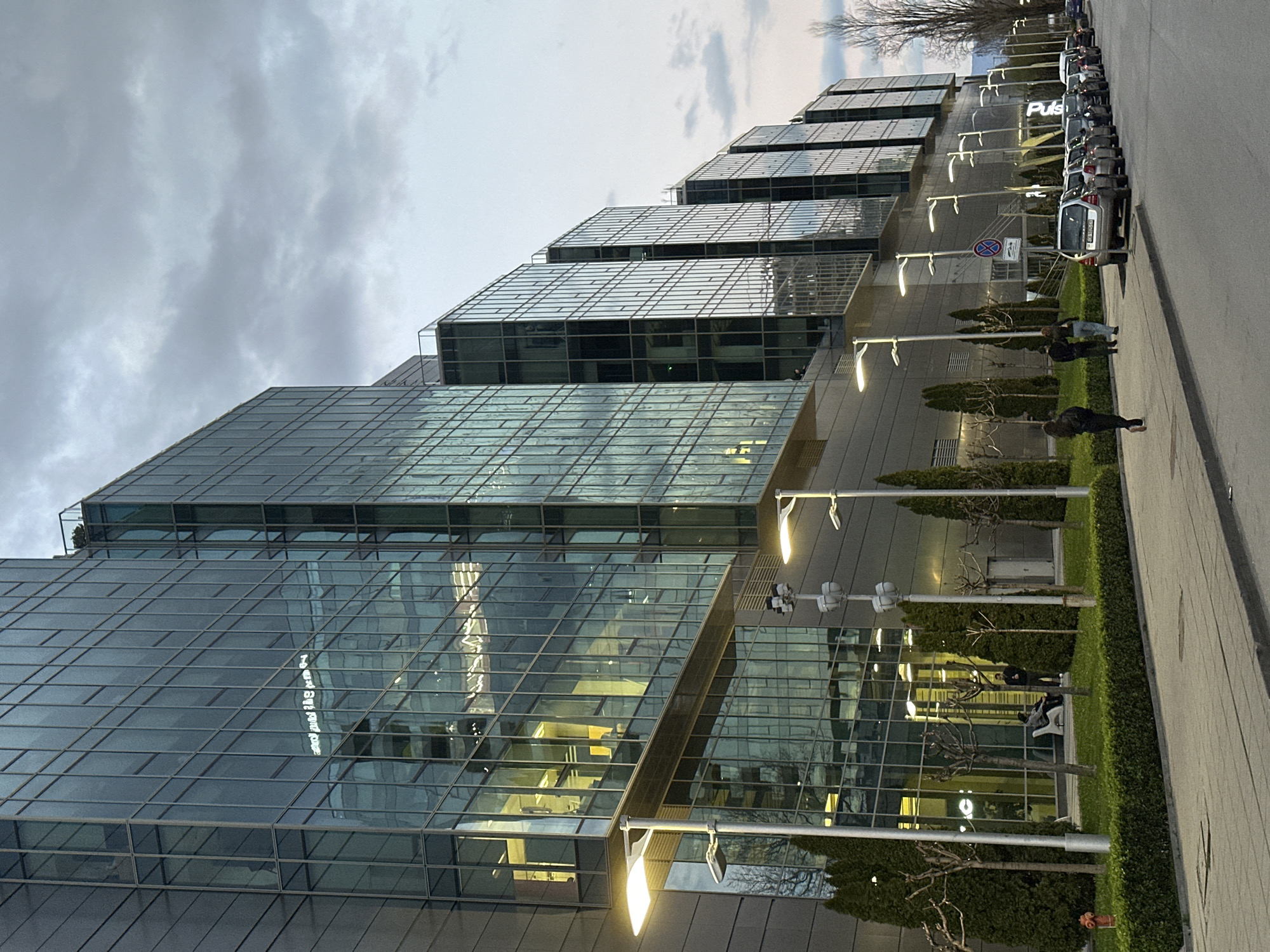 The image depicts a modern office building complex in Sofia, Bulgaria. The buildings are characterized by their glass facades and stepped design. The complex is likely located near a road, as evidenced by parked cars and pedestrian traffic. The sky is overcast, suggesting a cloudy day. People are walking along a paved path adjacent to the building. The location is possibly near the Sofia Airport, based on the style of the architecture.
