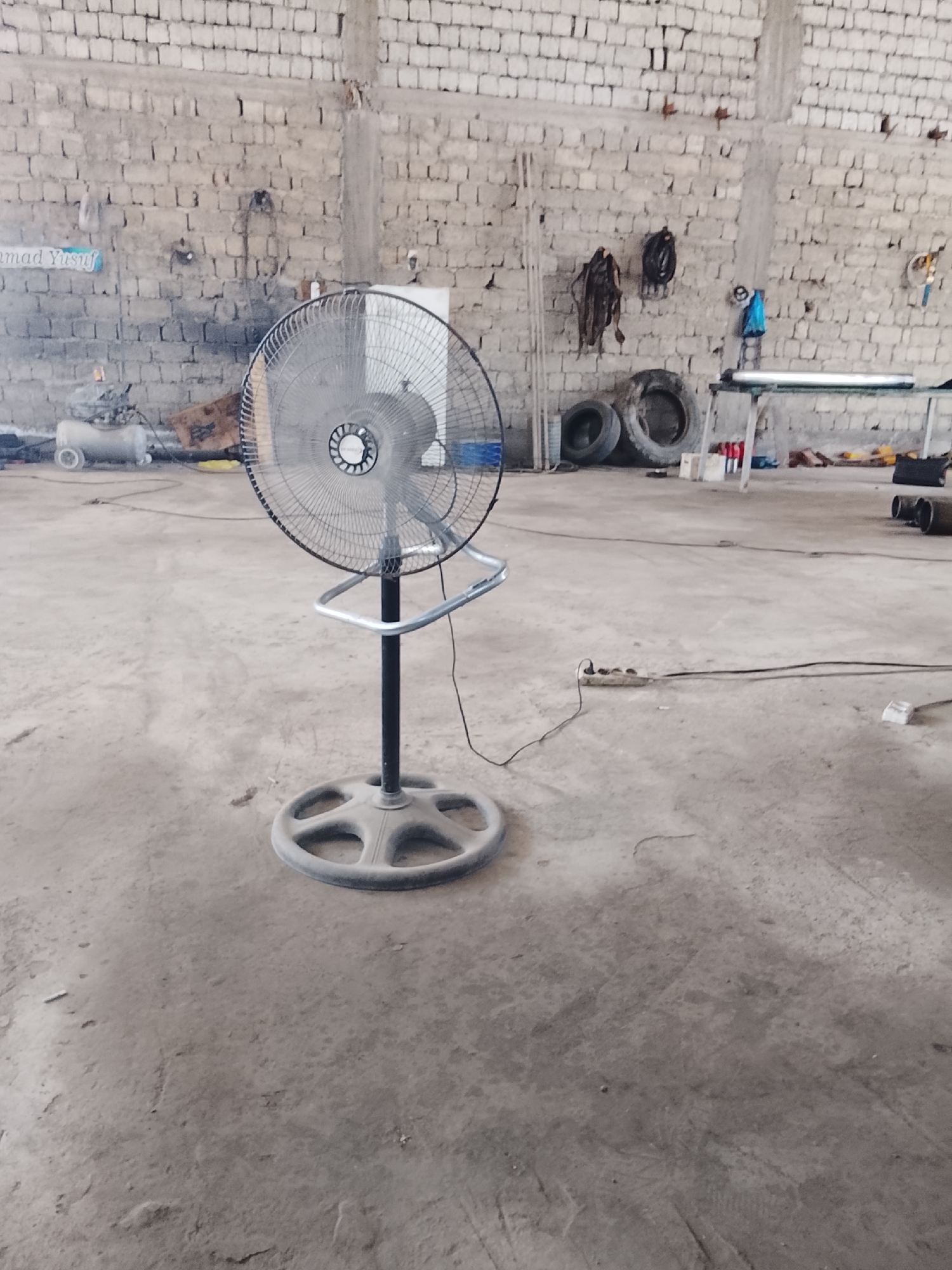 A stationary fan on a circular base stands in the center of a concrete floor, facing left. The fan's motor housing is visible, and the blades are obscured by a protective grill. Behind the fan, a brick wall stretches across the background, with various objects hanging on it, including what appear to be tools and electrical wires. To the left of the fan, a small air compressor with wheels is visible. The floor is dusty and uneven. The scene appears to be an industrial or workshop setting. The location is identified as Sabir Rakhimov, Uzbekistan.