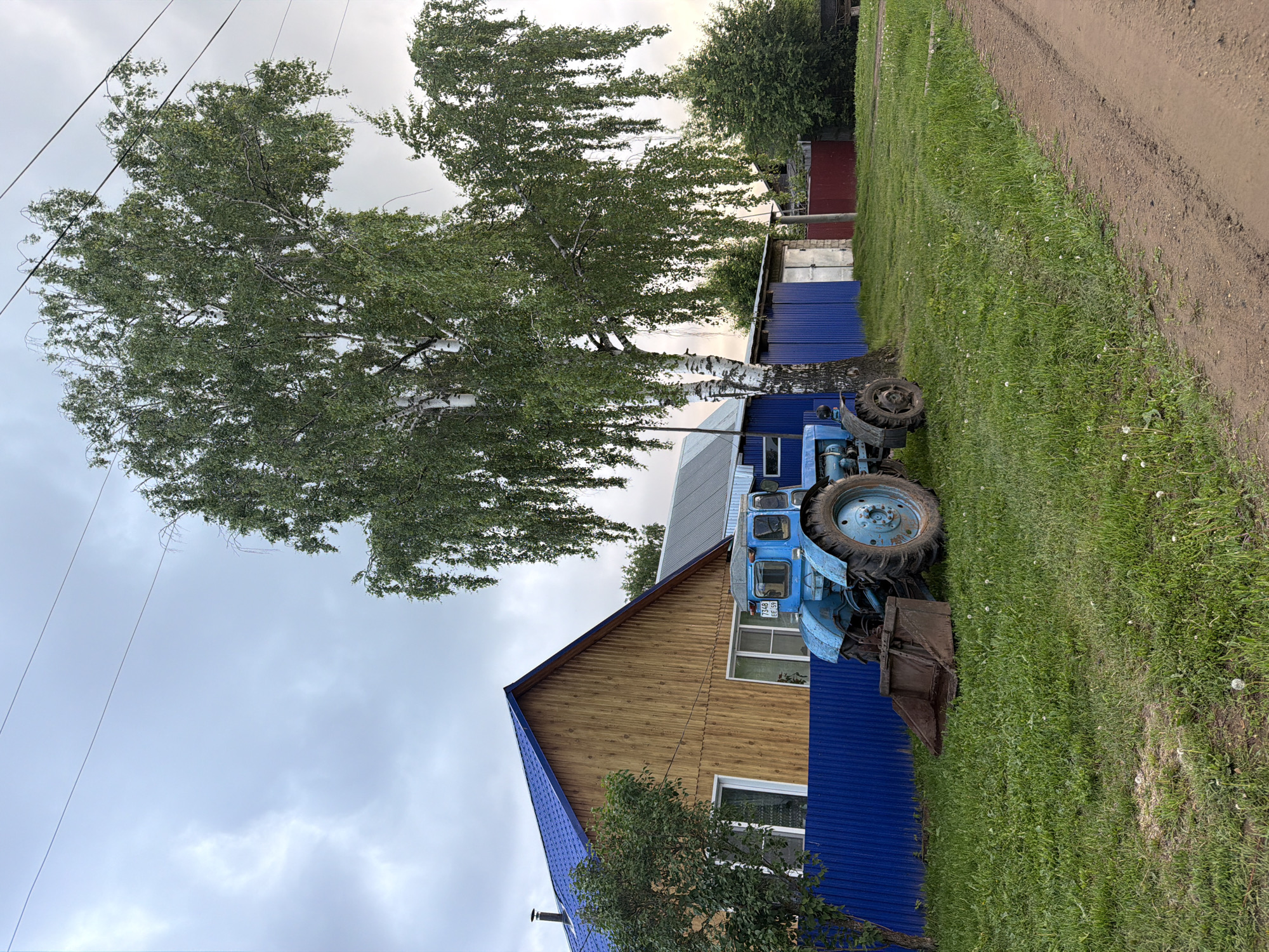 The image captures a rural or semi-rural scene in Marakushi, Russia, under an overcast sky. The primary subjects include a light blue agricultural tractor, a house, and a large birch tree.
The house, positioned in the midground, features a natural wood plank facade on its upper story and a vibrant blue corrugated metal roof, with a matching blue metal section forming part of its lower wall or foundation. Two windows are visible on the side of the house facing the viewer. Adjacent to the house, parked on a patch of green grass, is an older model light blue tractor, likely an agricultural type, equipped with a rusty front-end loader bucket. A tall, mature birch tree with distinctive white bark and dense green foliage stands prominently to the left of the house and tractor, its branches extending high into the cloudy sky.
To the right of the grass and tractor, a well-worn dirt road stretches into the distance. In the background, beyond the house and tractor, several smaller outbuildings with blue and red corrugated metal siding are partially visible, surrounded by additional trees and dense greenery. Overhead, several power lines traverse the cloudy, uniform gray sky.
No specific activity or event is taking place; the scene is static, depicting a moment in a rural landscape. The light suggests it is daytime under an overcast condition, providing soft, diffuse lighting without strong shadows. The visible text on the tractor's license plate reads "P348 BEE 159".