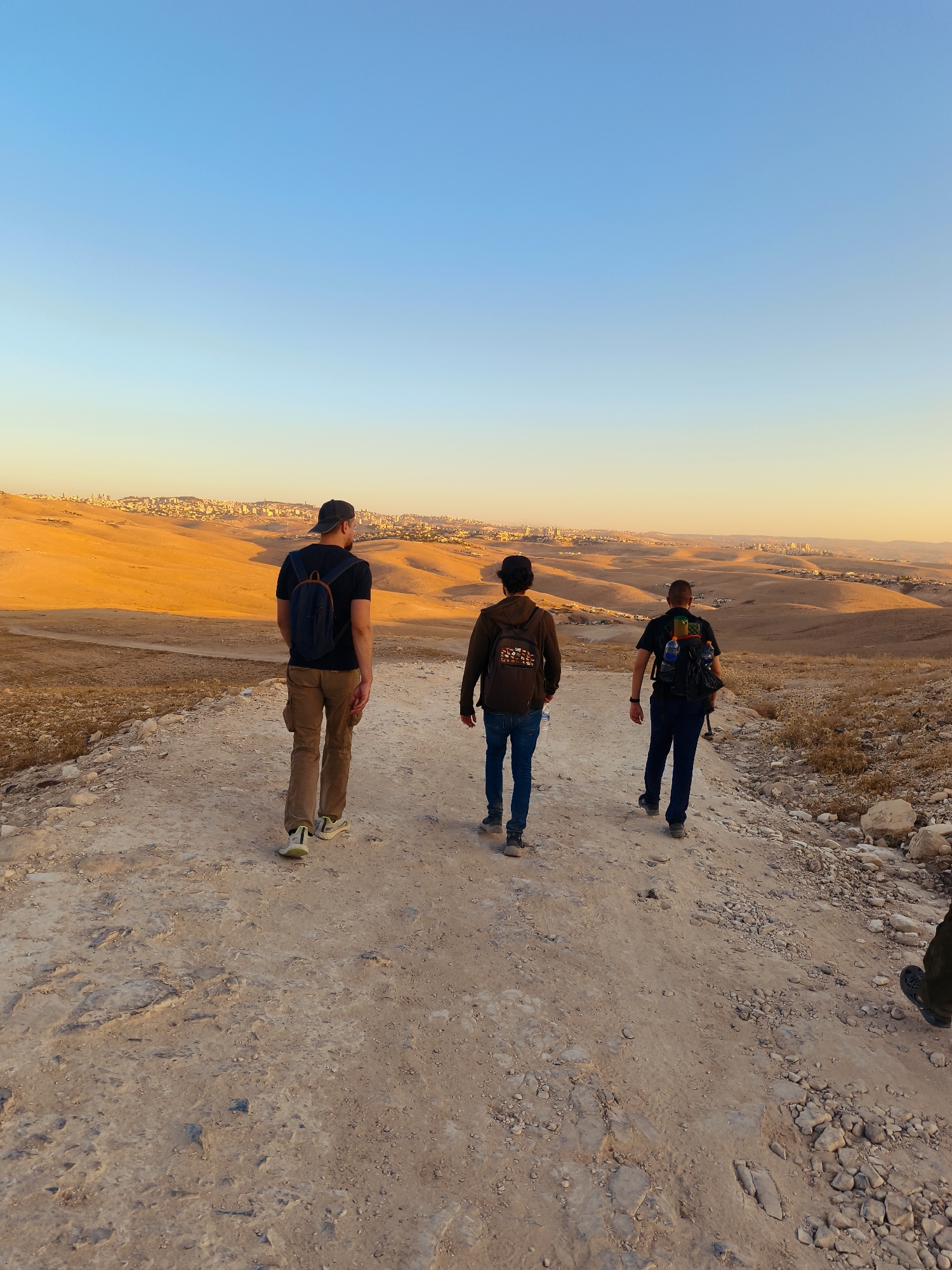 Three individuals are walking away from the camera on a dirt path in a desert landscape. The person in the center wears a black t-shirt and dark pants with a black backpack. To their left, a person in a black t-shirt and tan pants with a dark backpack and a baseball cap is walking. To the right of the center person, another individual in a dark shirt and pants with a black backpack is walking. They are in a dry, arid environment with rolling hills in the background, and a city is visible on the horizon, illuminated by the setting sun. The sky is clear and transitions from a light blue to a pale yellow near the horizon. The path is rocky and dusty.