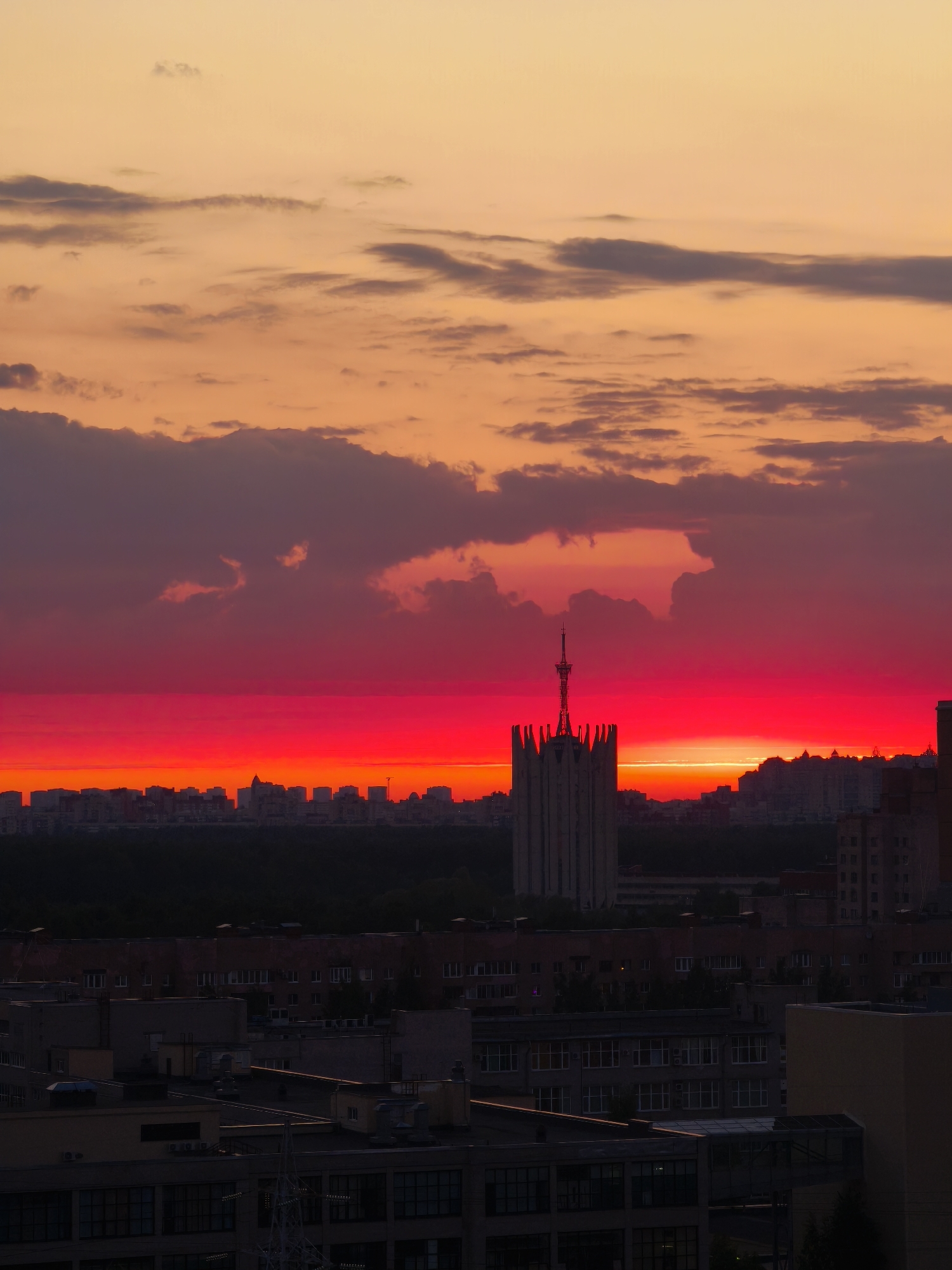The image captures a vibrant sunset over an urban landscape, likely Saint Petersburg, Russia. The sky transitions from orange at the top to a striking red horizon where the sun is setting. Dark clouds are interspersed within the red sky. In the foreground, a series of buildings dominate, including what appears to be an iconic Stalinist architecture building. The foreground buildings are darker, as the sun is setting.