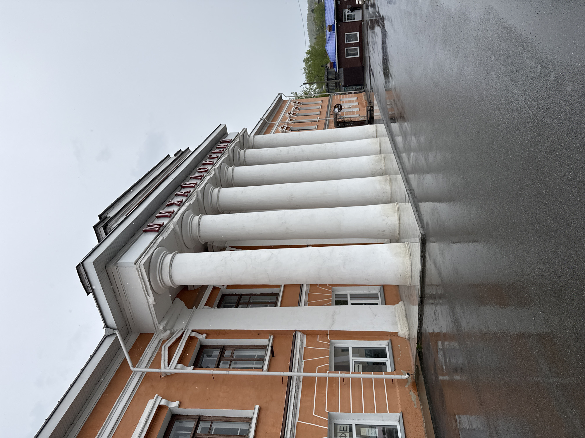 A low-angle, rotated view depicts the Mias Museum of Local Lore (Музей Миасский Краеведческий), in the city of Miass, Russia. The building's facade, painted orange, features several tall, white cylindrical columns under a white roof. Windows are visible on the building’s facade. The sky is overcast. Wet pavement is visible in the foreground. A second building can be seen in the distance.
