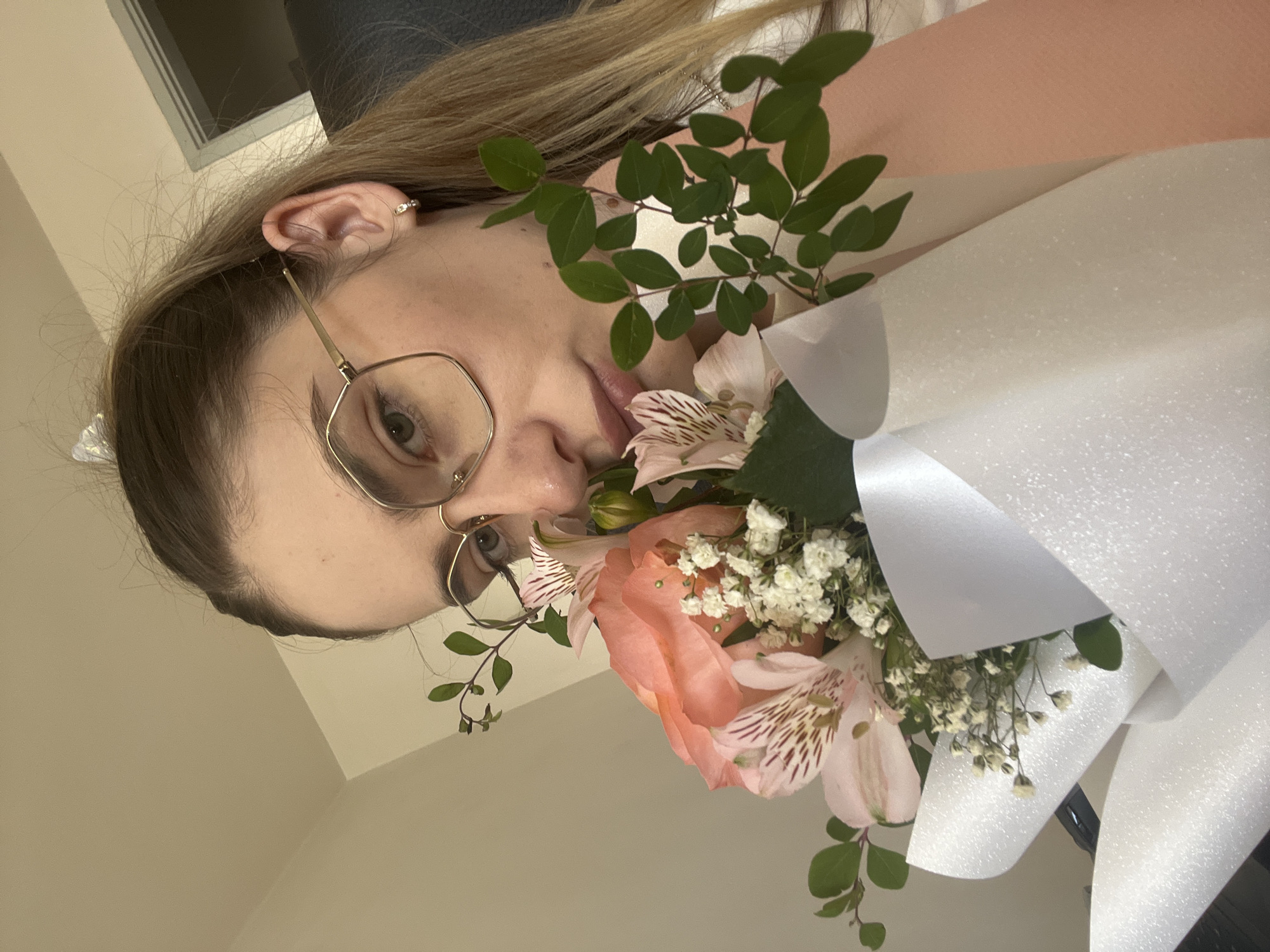 A medium close-up shot shows a young woman with blonde hair, wearing gold-rimmed glasses and small hoop earrings, holding a bouquet of pink and orange flowers, along with greenery and small white flowers. The woman is looking at the camera with a neutral expression. She is wearing a white top. The bouquet is wrapped in white and pink paper. The background is a plain, light-colored wall, with a door frame visible in the upper right corner. The lighting is soft and diffused, suggesting an indoor setting.