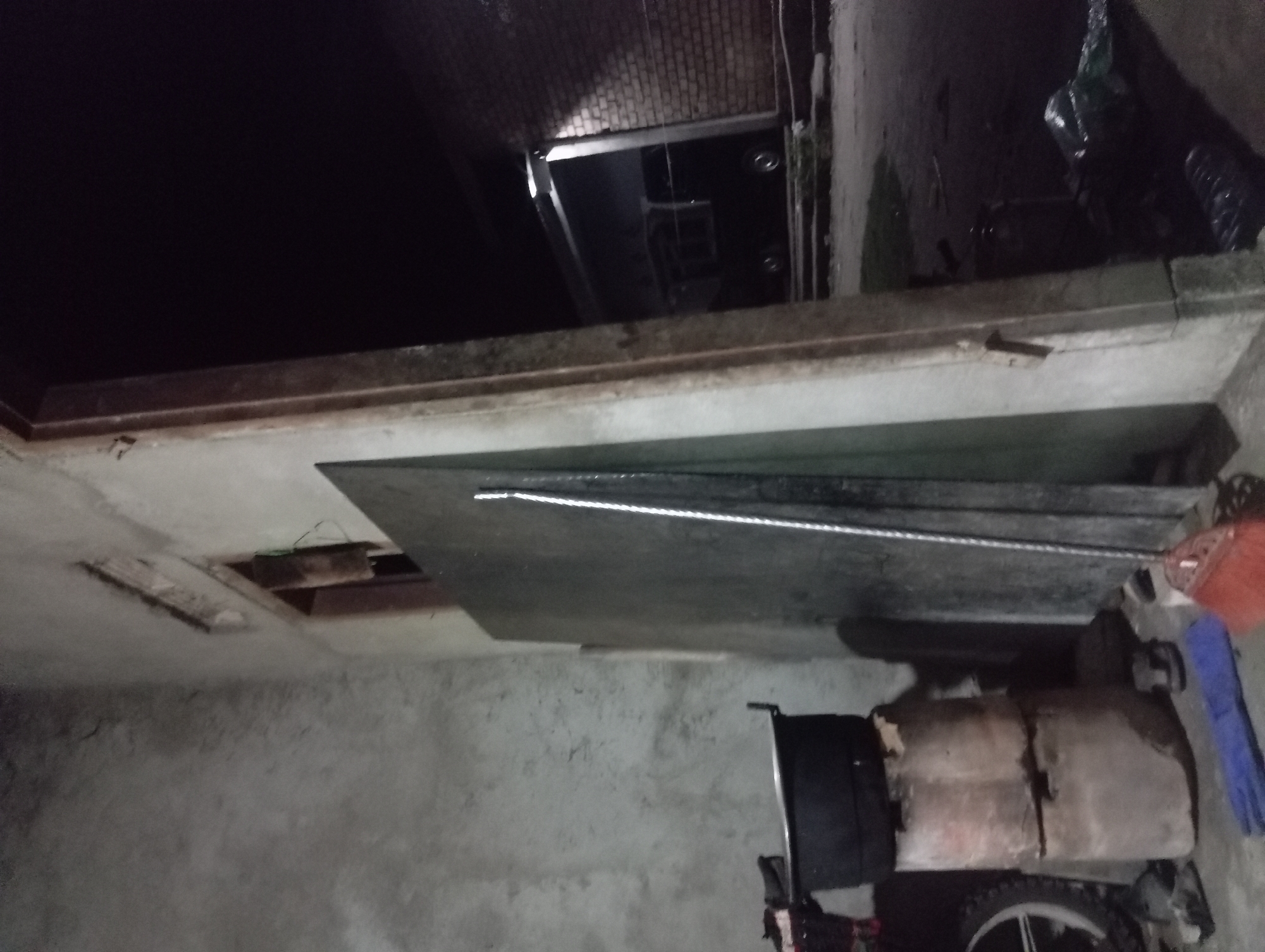 The image depicts a dimly lit, outdoor scene, possibly a backyard or alleyway, located in Rejosari, Indonesia. A large, dark metal pot rests atop two stacked concrete blocks, which are weathered and cracked. To the right of the pot, a tall, thin object, appearing to be a metal rod or pole, leans against a large, flat, dark gray panel. In the foreground, a red-bristled broom with a metal handle lies on the ground. Adjacent to the broom is a folded blue cloth. Further to the right, a section of a brick wall is visible, and in the background, through an opening or doorway, a dark-colored vehicle with visible wheels is parked. The ground appears to be concrete or dirt. The overall impression is one of a utilitarian, possibly neglected, outdoor storage area.