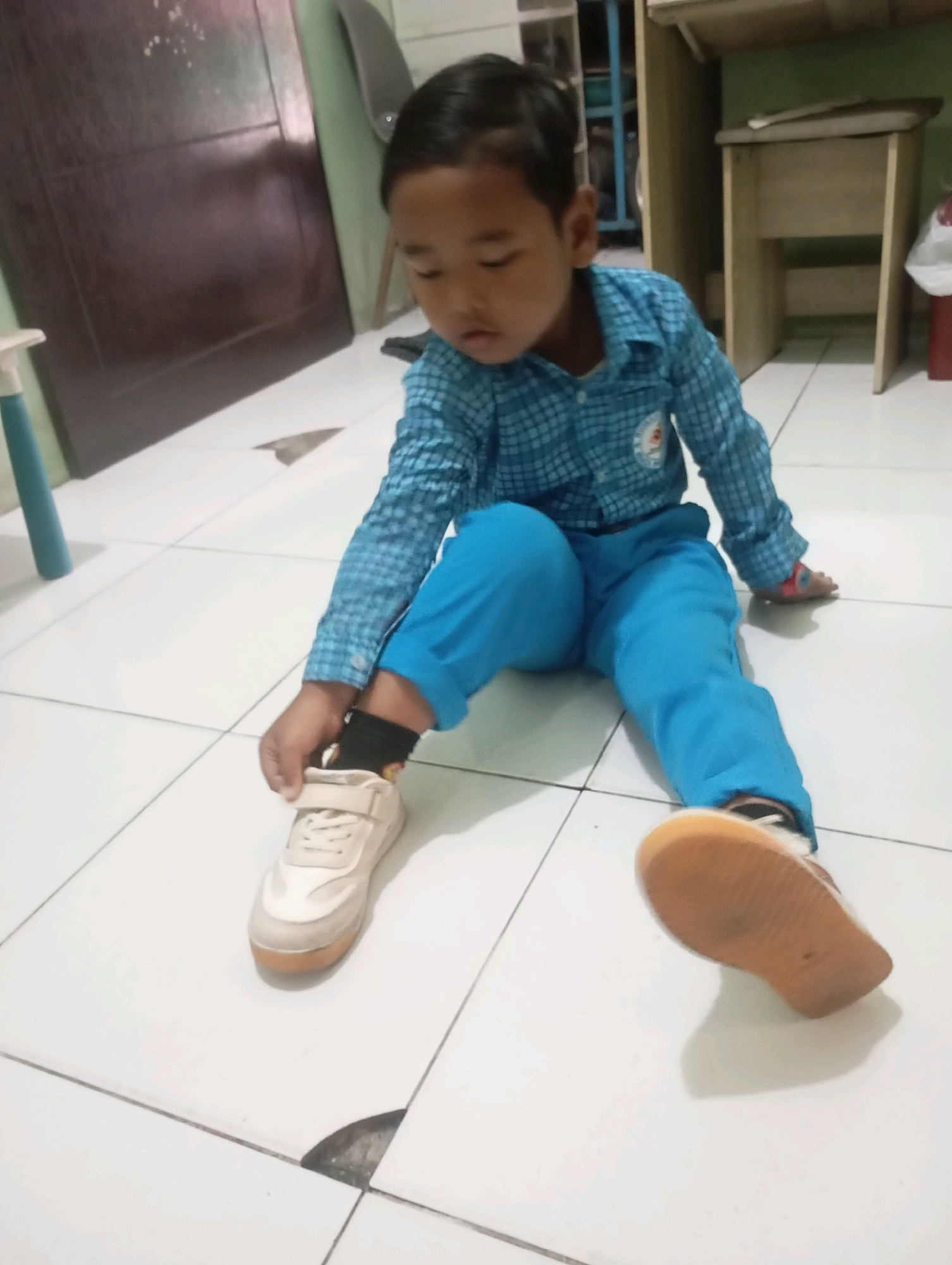 A young child, appearing to be of East Asian descent, is sitting on a tiled floor in what appears to be an indoor setting. The child is wearing a blue checkered long-sleeved shirt and bright blue pants. One foot, with a black sock on, is being adjusted with the child's right hand, and a light-colored sneaker with a velcro strap is visible. The other foot is partially obscured but also has a sneaker on. In the background, there is a wooden desk or table, a dark brown door or cabinet, and a partially visible green wall. The lighting suggests an indoor environment, possibly a home or classroom. The image is taken from a slightly elevated angle, focusing on the child. There is no discernible indication of the city of Babelan, Indonesia, in the visual information provided.