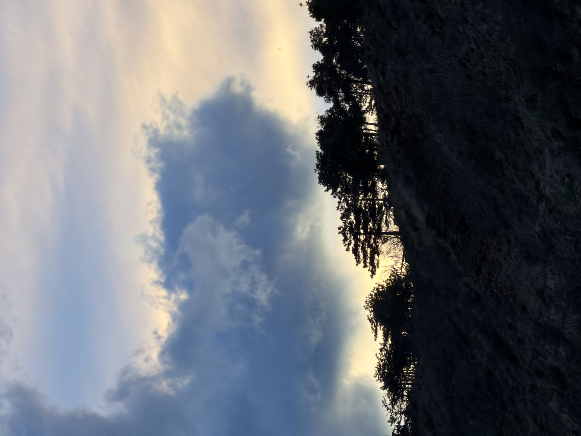 The image shows a sky with clouds illuminated by the setting sun. The clouds range in color from a light yellow-white near the sun to shades of blue and gray. On the right side of the image, there is a dark, silhouetted cliff face with sparse vegetation. The overall orientation of the photograph suggests it was taken looking upward at an angle. There is no direct way to determine that the location is Tbilisi, Georgia.