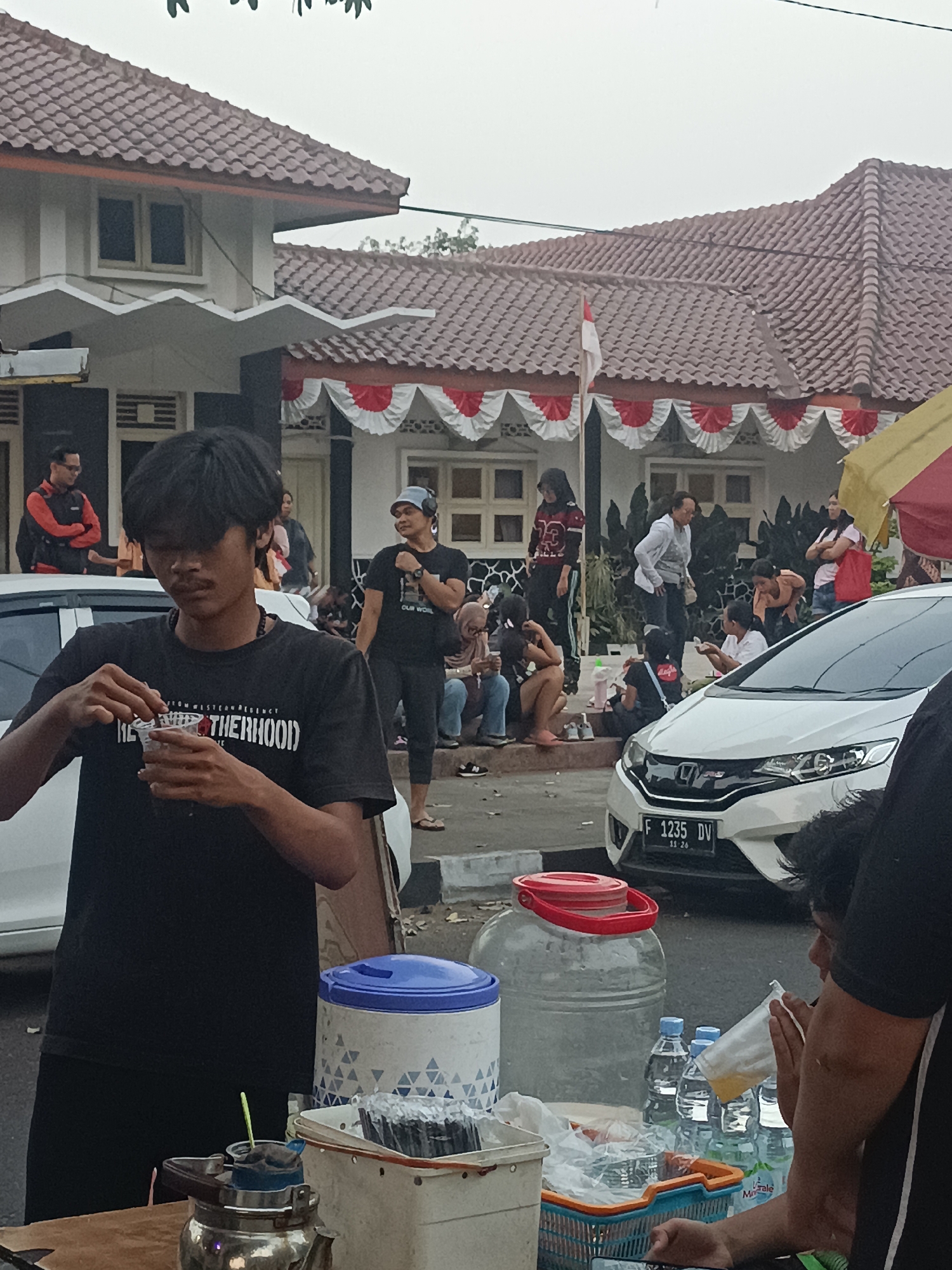 In a street scene in Sleman, Indonesia, on an overcast day, a young man with dark hair is the central subject, holding a small plastic cup and a stirrer in his hands, appearing to prepare a drink. He wears a black t-shirt with white text that reads "FROM WESTERN REGENCY BROTHERHOOD." To his right, a large transparent water dispenser with a red lid sits on a table. In front of the dispenser are several plastic water bottles, one of which is being held by another person whose arm and torso are visible on the far right.
In the background, several people are gathered. A white car, a Honda Jazz with the license plate "F 1235 DV," is parked in the middle ground. Behind the car, a building with a red-tiled roof is decorated with red and white banners, suggesting a celebratory event or national holiday. The building has white walls and several windows. More people are visible in front of the building and on the sidewalk. A yellow and red umbrella is partially visible on the right side of the frame. The ground appears to be asphalt or concrete.