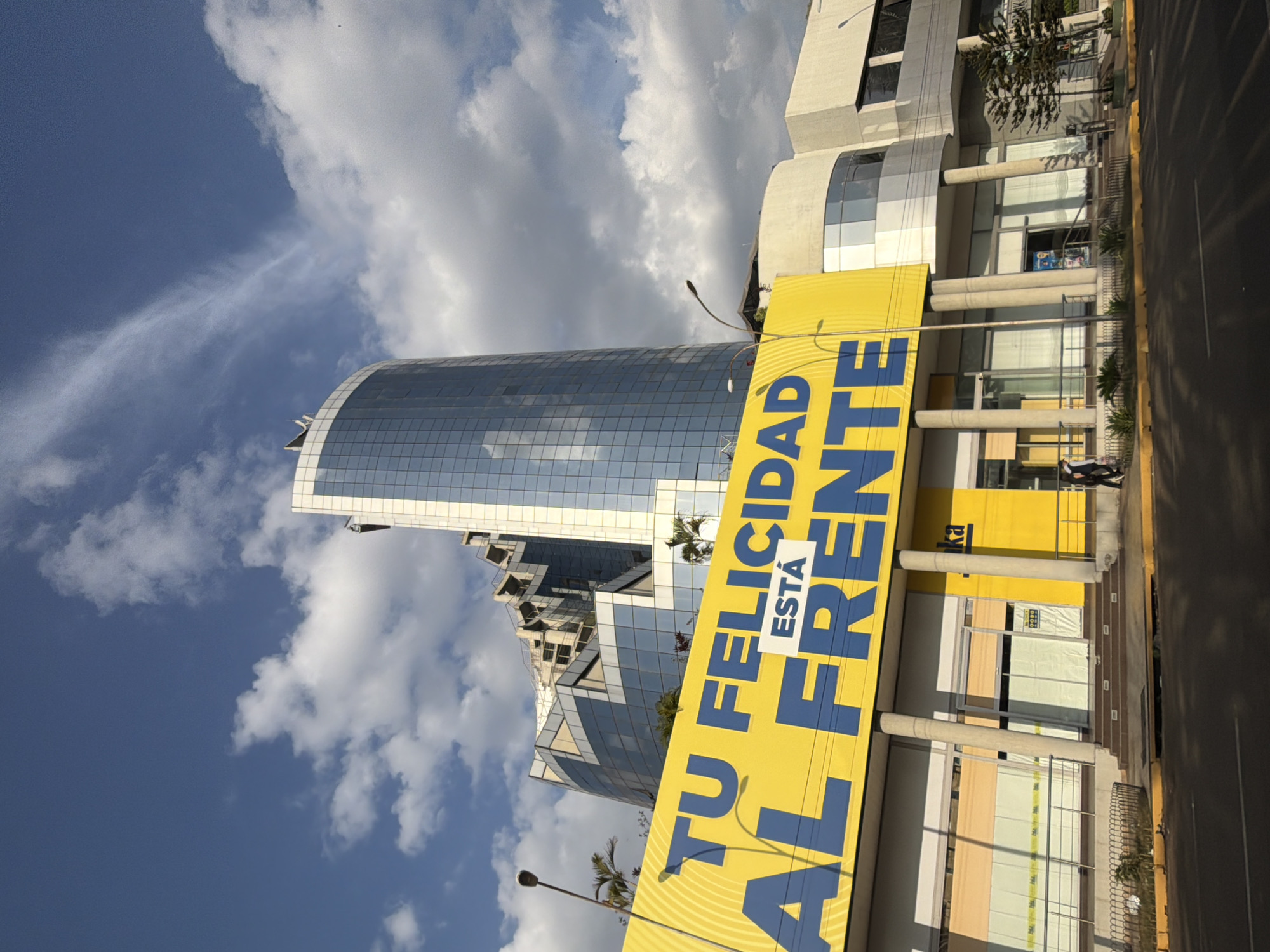 The image shows a street view with a tall, curved, glass-covered building in Ciudad Guayana, Venezuela. In front of the building, there's a large yellow banner with blue letters that read "TU FELICIDAD ESTÁ AL FRENTE." The sky is partly cloudy. There is a building with yellow accents adjacent to the banner. There is one visible pedestrian on the sidewalk next to the street. A street lamp is visible. Some greenery lines the sidewalk.