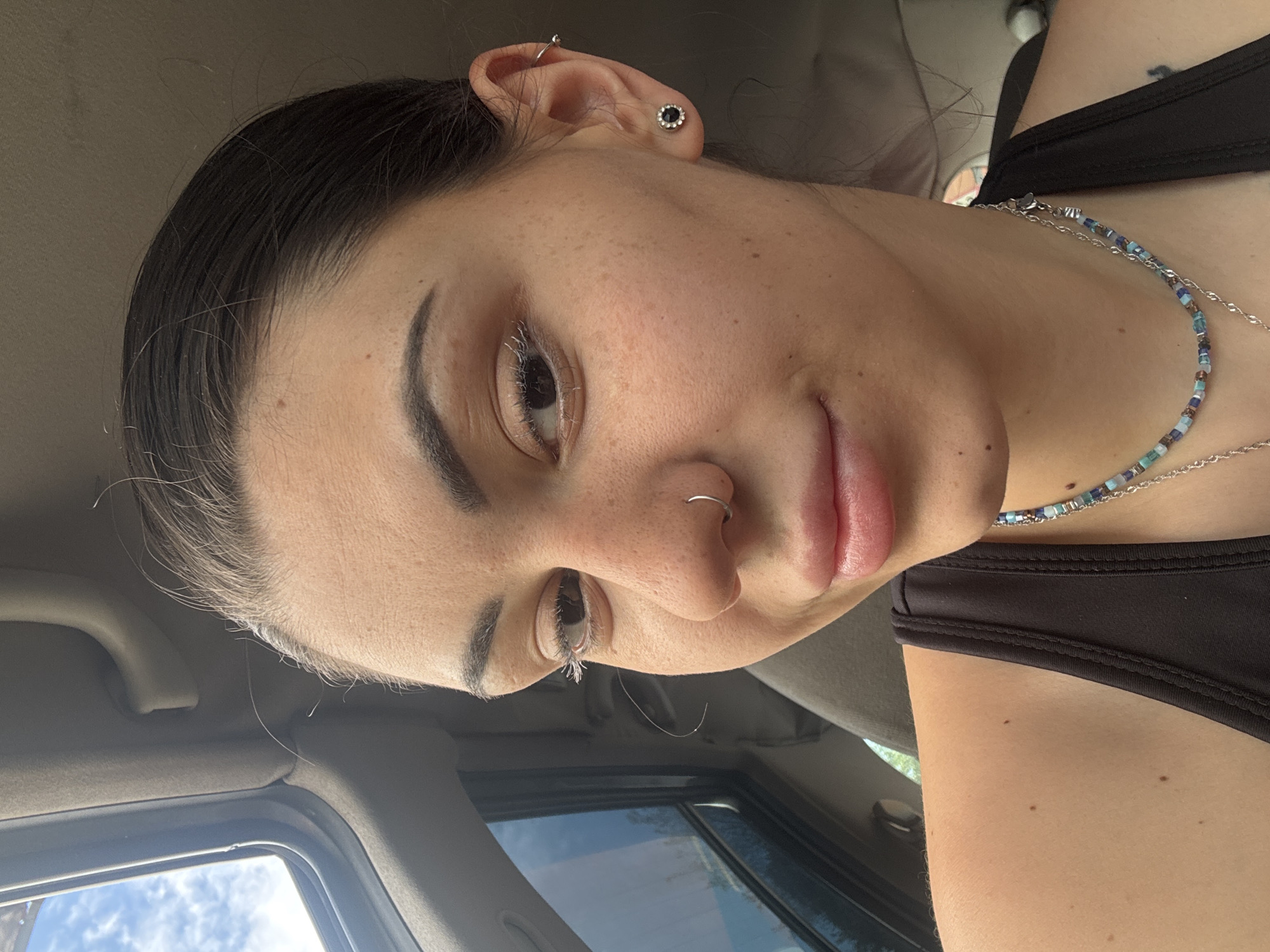 A close-up, tilted portrait features a young woman looking slightly upward and to the left. Her dark hair is pulled back, with some loose strands around her hairline. She has distinct eyebrows and a visible left eye with dark eyelashes. A silver hoop nose ring adorns her left nostril, and her lips are full and slightly parted in a faint smile.
Her skin shows numerous freckles across her face and visible moles on her skin. On her visible left ear, she wears two piercings: a black stud earring with a silver rim in the lobe and a silver hoop helix piercing. She is dressed in a black top or swimsuit. Around her neck, she wears a beaded necklace with small blue, green, and brown square beads, layered with a delicate silver chain.
The background elements, including the car's headliner, window frame, and a clear view of a bright, cloudy sky through the window, indicate she is inside a vehicle during daytime. The lighting is natural, highlighting her face. No text is visible in the image.