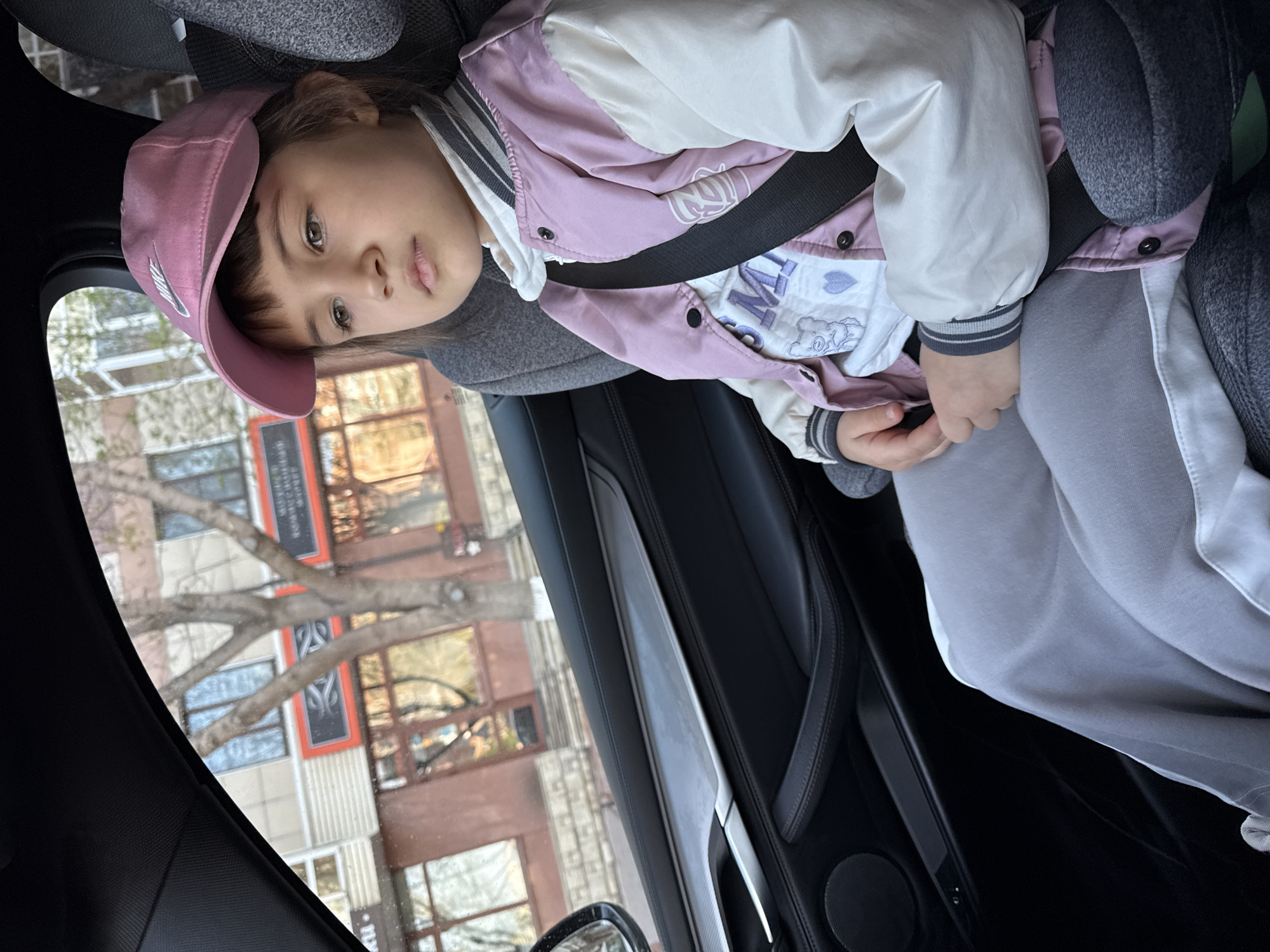 A child is seated in the passenger seat of a car, wearing a pink baseball cap and a jacket with a white and pink color scheme. The child is secured with a seatbelt. Outside the car window, a building is visible, which is located at 122 Mira Avenue in Krasnoyarsk, Russia. A bare tree is also visible outside the car.