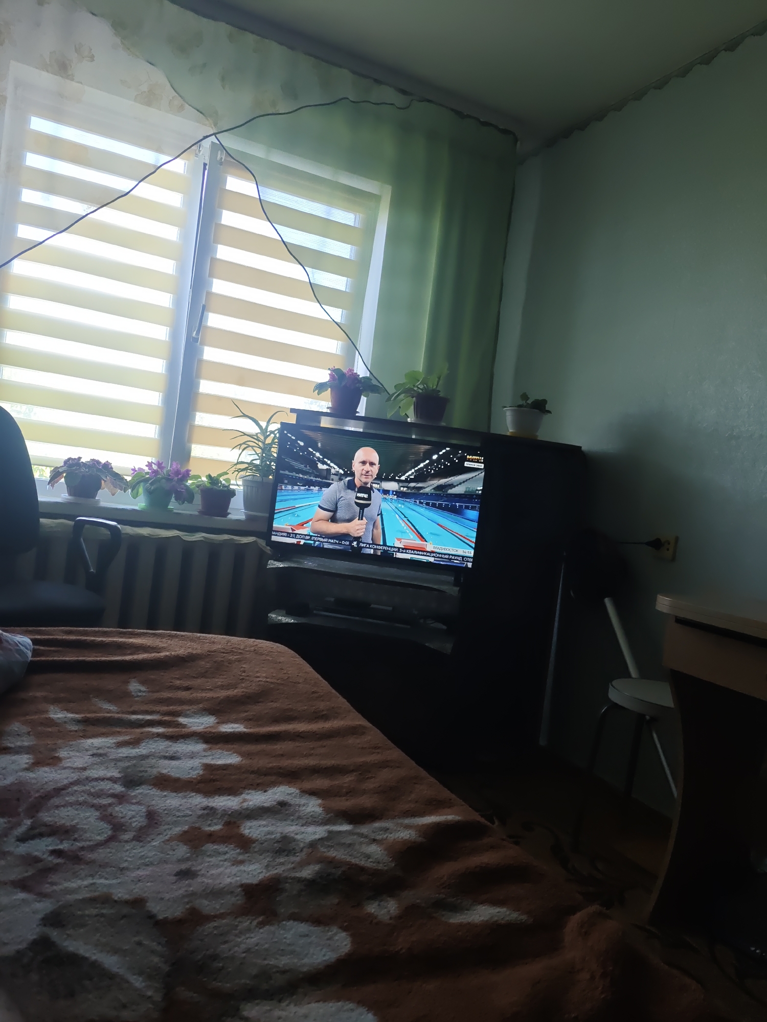 In a dimly lit room, likely a bedroom, a television displays a man holding a microphone, standing in front of a swimming pool. The TV is placed on a dark wooden cabinet. A window with horizontal blinds is visible on the left, with several potted plants on the windowsill. The room appears sparsely furnished, with a bed covered by a brown blanket with a floral pattern in the foreground. A dark office chair is positioned next to the bed. The walls are painted a light green color, and a section of the ceiling shows some disrepair. Electrical wires are visible near the ceiling. The scene suggests a domestic setting in a city like Курашим, Russia, where everyday activities like watching television are taking place.