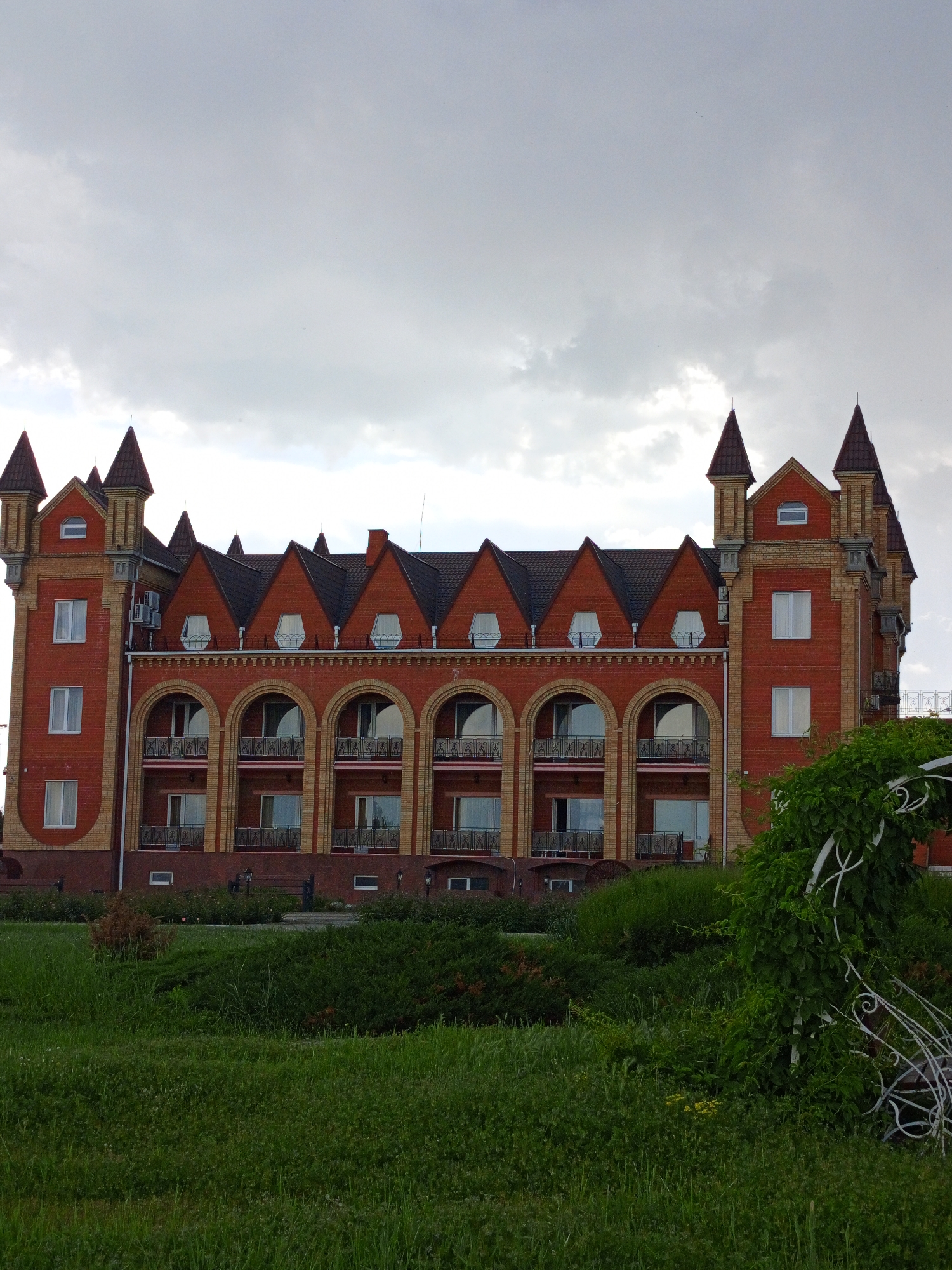 The image shows a large, two-story building with a reddish-brown brick exterior. The architectural style suggests a castle or fortress, with turret-like structures on the corners and pointed roofs along the top. Each window on the second story has a curved arch above it, and there are small balconies visible. The ground in front of the building is covered in green grass and shrubbery, creating a lawn-like setting. The sky above is cloudy and overcast. The location is Unknown, Unknown.