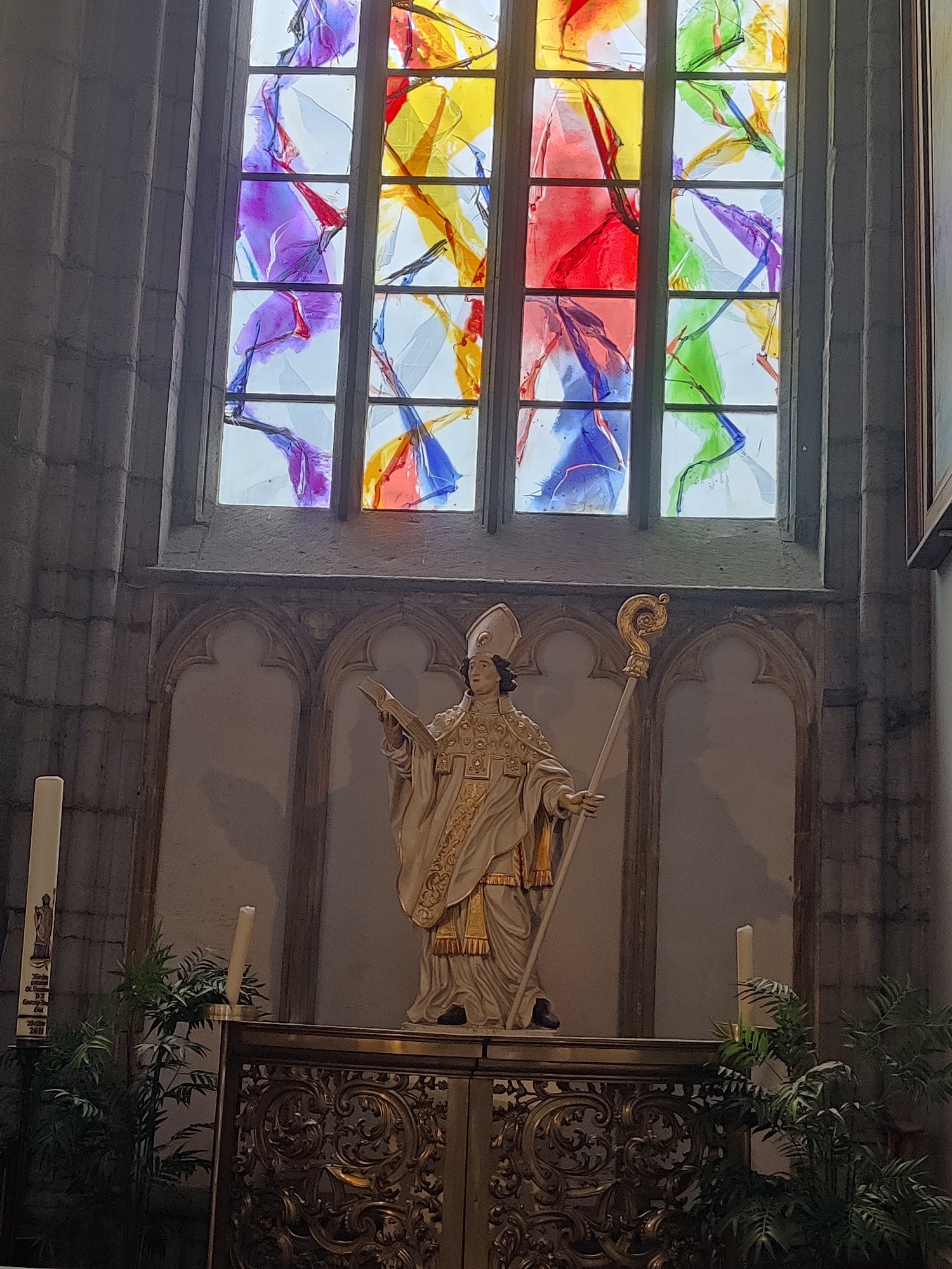 The image depicts the interior of a church, specifically the chancel area, featuring a stained-glass window, a statue, and an altar. The stained-glass window is large and abstract, with irregular shapes and vibrant colors including purple, red, yellow, green, and blue, set within a grid of leaded panes. Below the window, a statue of a religious figure, likely a saint or bishop, stands prominently. The figure is depicted in ornate ecclesiastical robes, wearing a mitre and holding a crozier in its right hand and a book in its left. The statue is positioned behind a decorative altar or communion rail, which is ornate and made of metal with intricate scrollwork. Two tall, white candles are visible: one to the left of the statue and another to the right, with a potted plant placed in front of the altar on the right side. The wall behind the statue is adorned with Gothic-style niches. The architecture suggests a historic European church. The scene is likely located in Liège, Belgium, within a church dedicated to Saint Lambert, as indicated by the presence of his statue and the significance of the saint in the city's history.