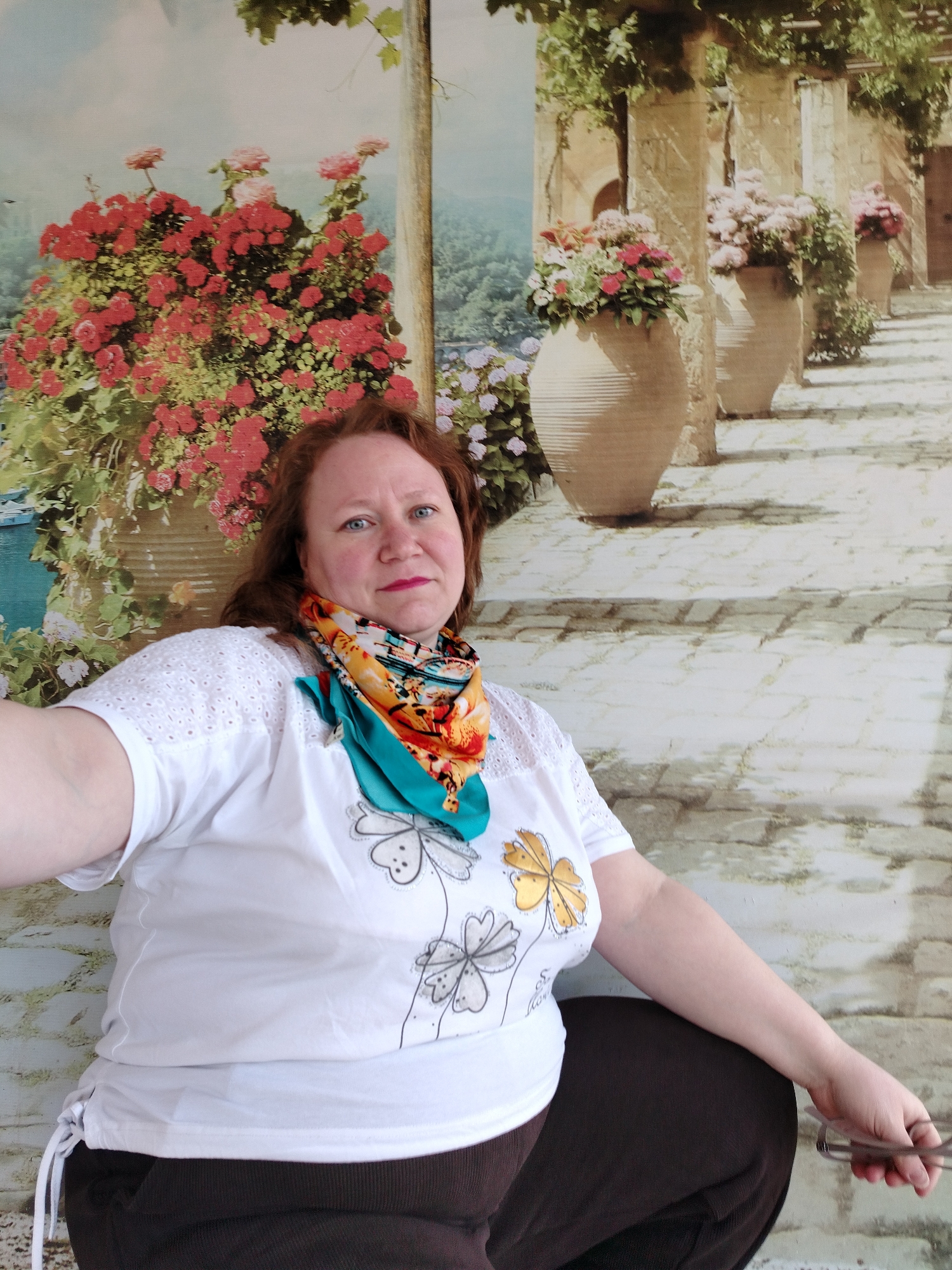 A medium shot depicts a middle-aged Caucasian woman with reddish-brown hair and blue eyes. She is wearing a white T-shirt with flower designs and a multicolored scarf. The woman is seated and appears to be holding eyeglasses in her right hand. The background showcases a wall or backdrop with a painted scene of flowers and a path leading to large vases filled with more flowers. The location is identified as Chaikovsky, Russia.