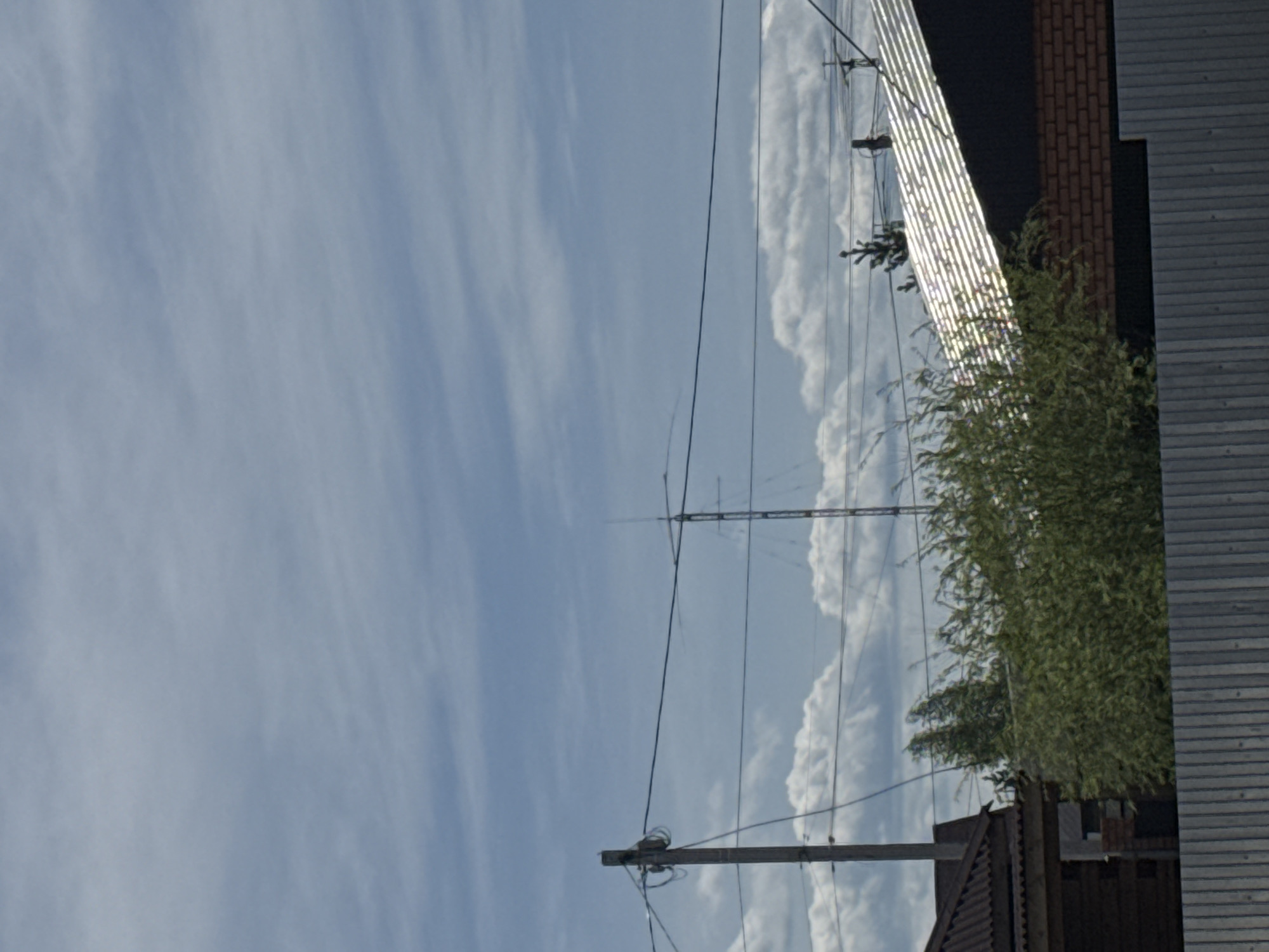 The image captures a sky with scattered clouds. A radio tower stands vertically in the scene, bisected by multiple horizontal elements. A utility pole with wiring is visible at the bottom of the frame. The right side of the image features a building with mixed materials, including siding, brick, and reflective panels, partially obscured by greenery. The image is rotated 90 degrees counterclockwise. The location appears to be residential or suburban, but its exact whereabouts within the city of Unknown, Unknown is unknown.